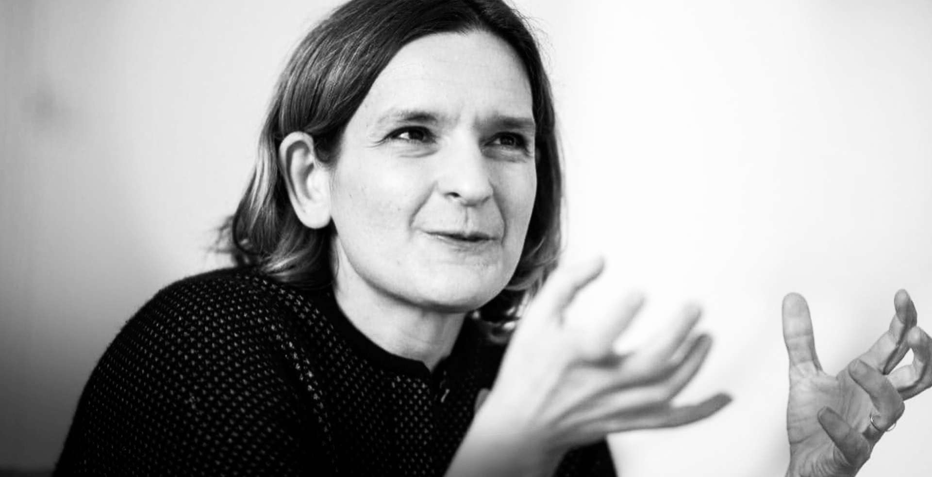 A black-and-white photograph of Esther Duflo with shoulder-length hair, gesturing with her hands while speaking.