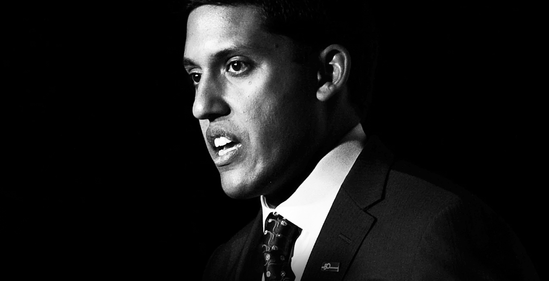 Black and white portrait of Dr. Rajiv Shah in a suit, facing left with mouth slightly open, against a black background.
