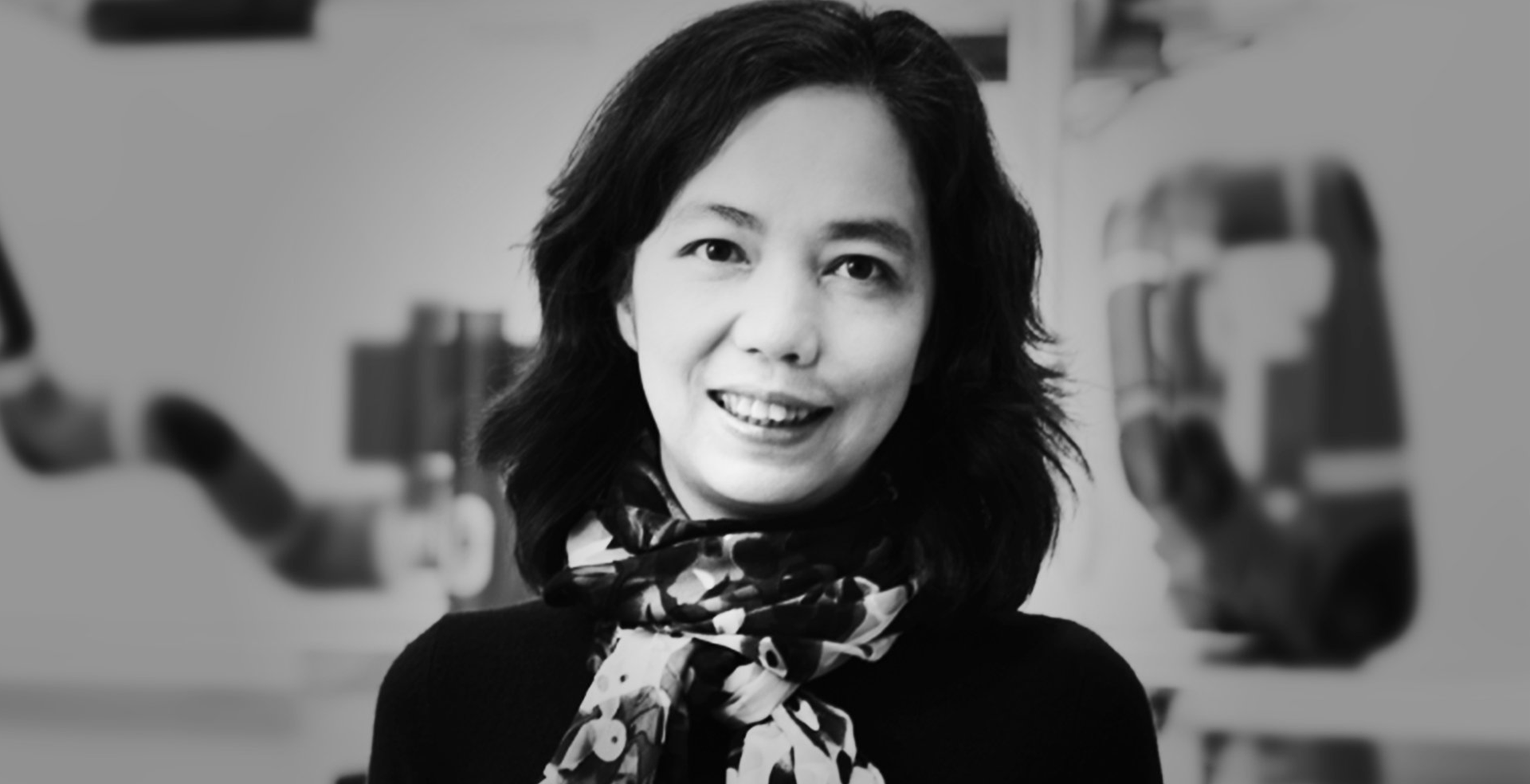 Black and white photo of Dr. Fei-Fei Li, smiling at the camera, pointing at it with both hands. She is wearing a patterned scarf and a dark top, with a blurred indoor background.