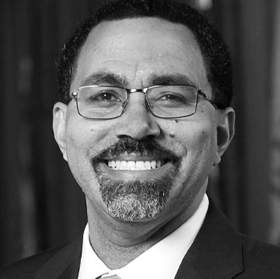 Black and white portrait of a smiling John King with glasses, a beard, and wearing a suit and tie.