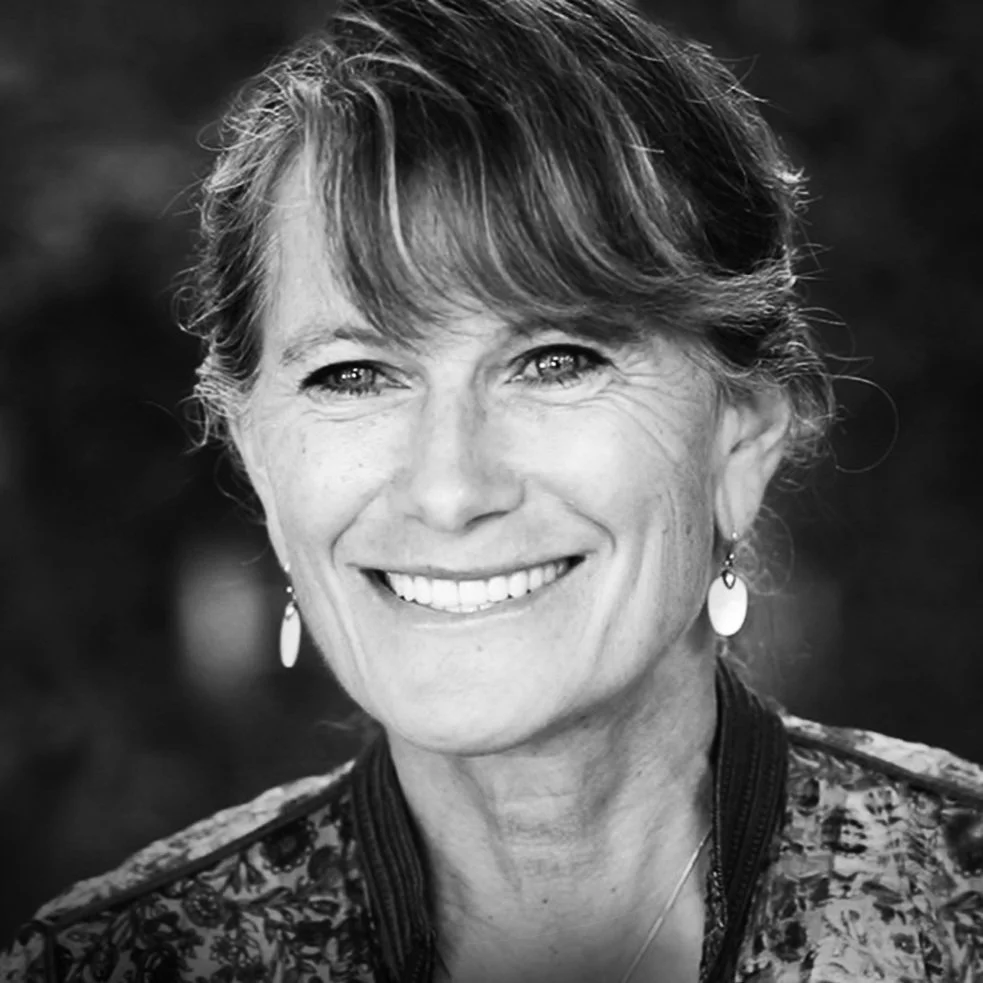 Black and white photo of a smiling Jacqueline Novogratz, wearing earrings and a patterned jacket.
