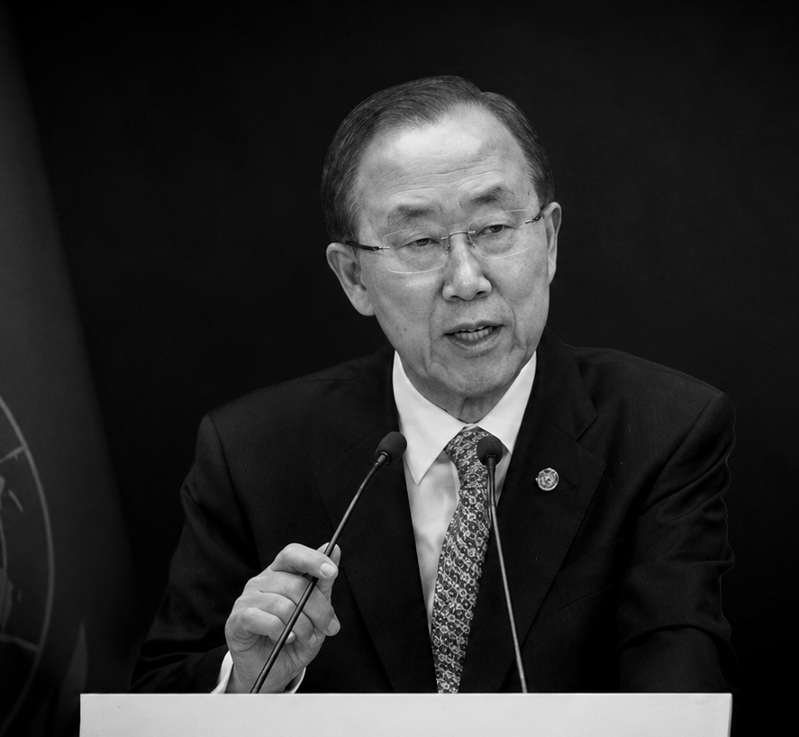 Black and white photo of Ban Ki-Moon speaking at a podium with two microphones, wearing glasses, a suit, and a patterned tie.