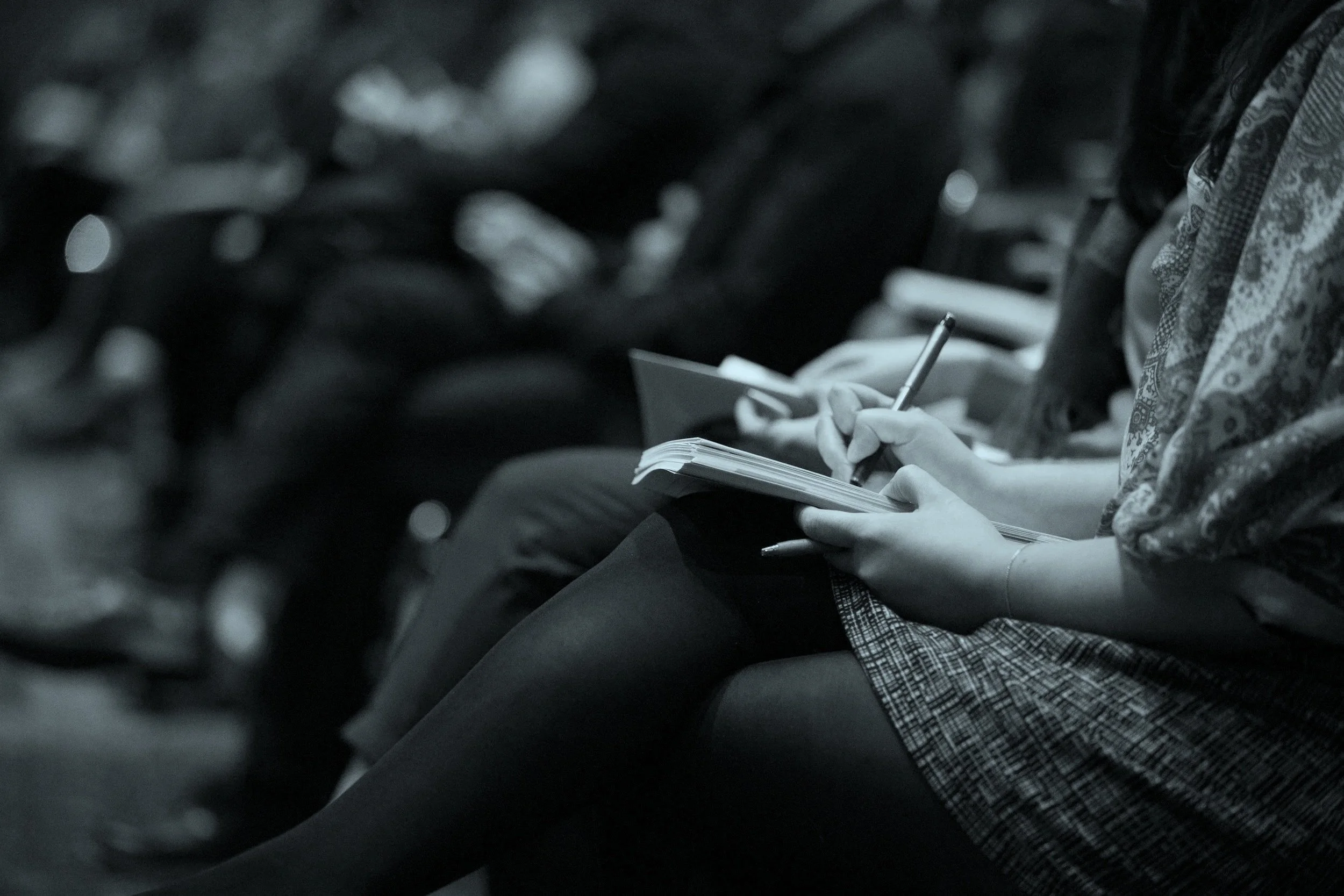 A person sitting on a bus or train, writing in a notebook with a pen. They are wearing a patterned jacket or scarf and have a watch on their wrist. A classroom image, representing the delivery of cyber security awareness training.