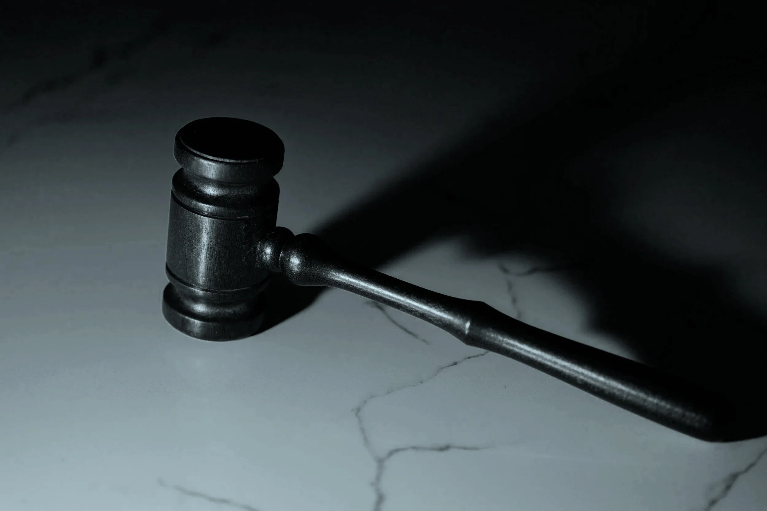 Close-up of a black gavel resting on a white surface with faint cracks, casting a shadow in a dimly lit setting. An image which represents regulatory compliance in Cyber Security.