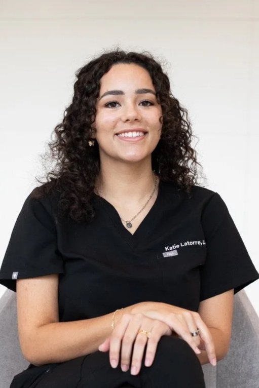 A woman with curly dark hair wearing a black uniform, sitting with her hands folded on her lap, smiling, with a white background.