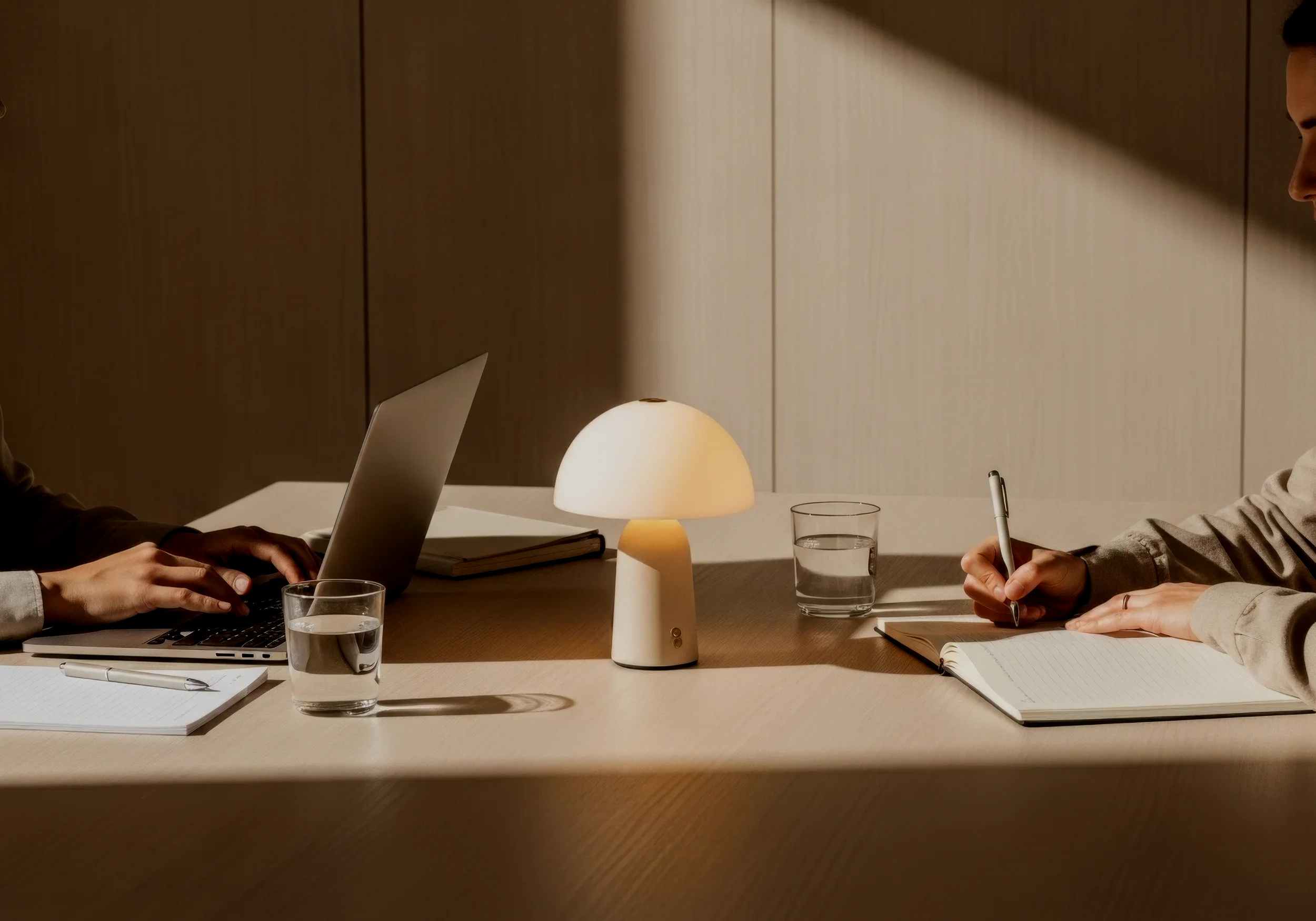 People sitting at a table having a meeting, with a lamp, notebooks, glasses of water, and a laptop in a modern office setting.