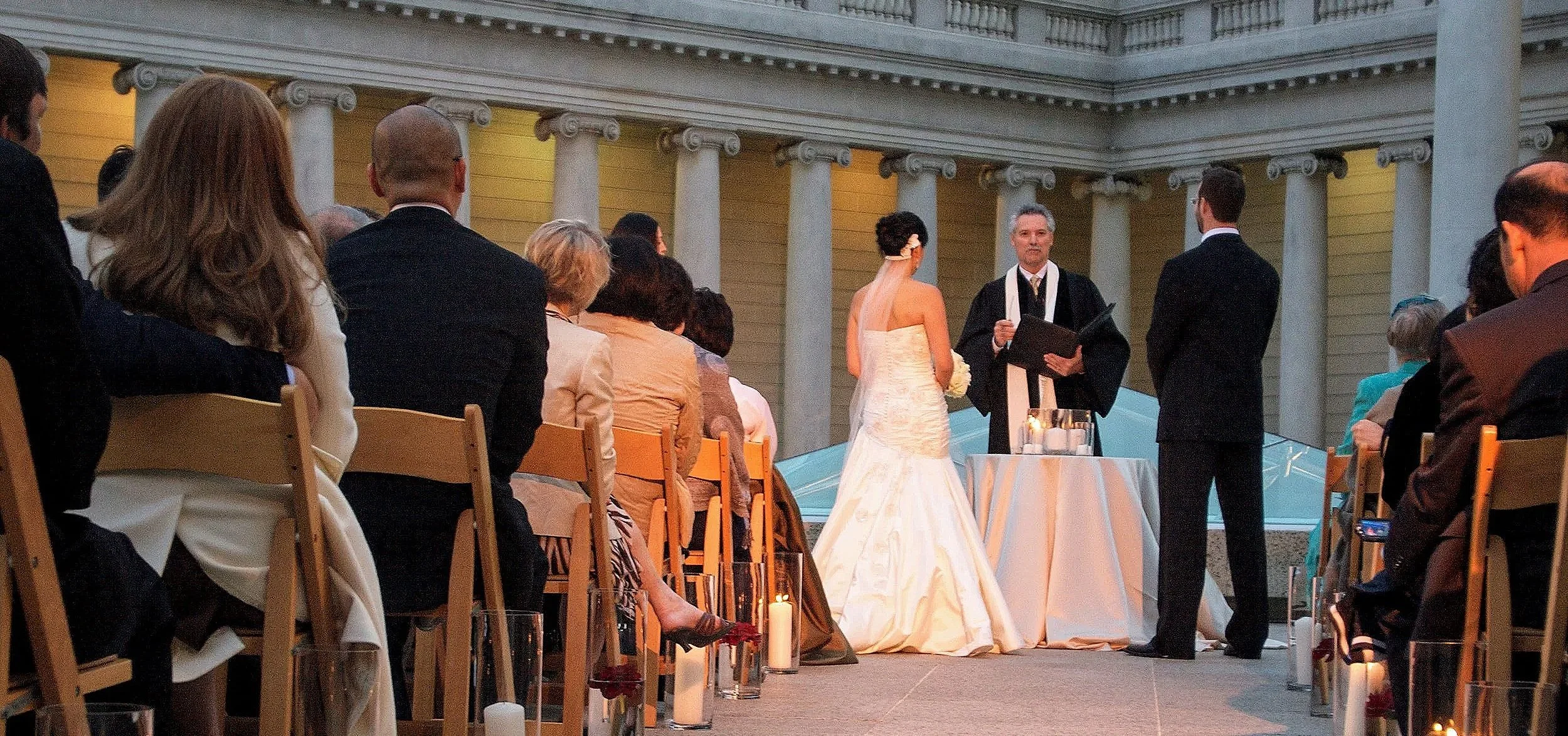 A wedding ceremony taking place outdoors at sunset, with the bride and groom exchanging vows in front of seated guests, surrounded by candles and decorative elements.