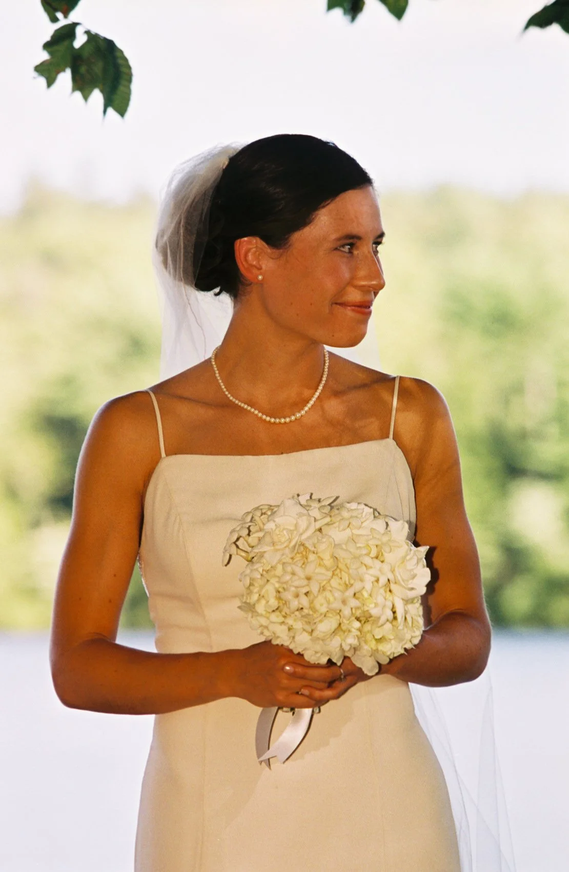 A bride in a white wedding dress holding a bouquet of white flowers, with dark hair styled in an updo and wearing a pearl necklace and earrings, standing outdoors with green trees blurred in the background.