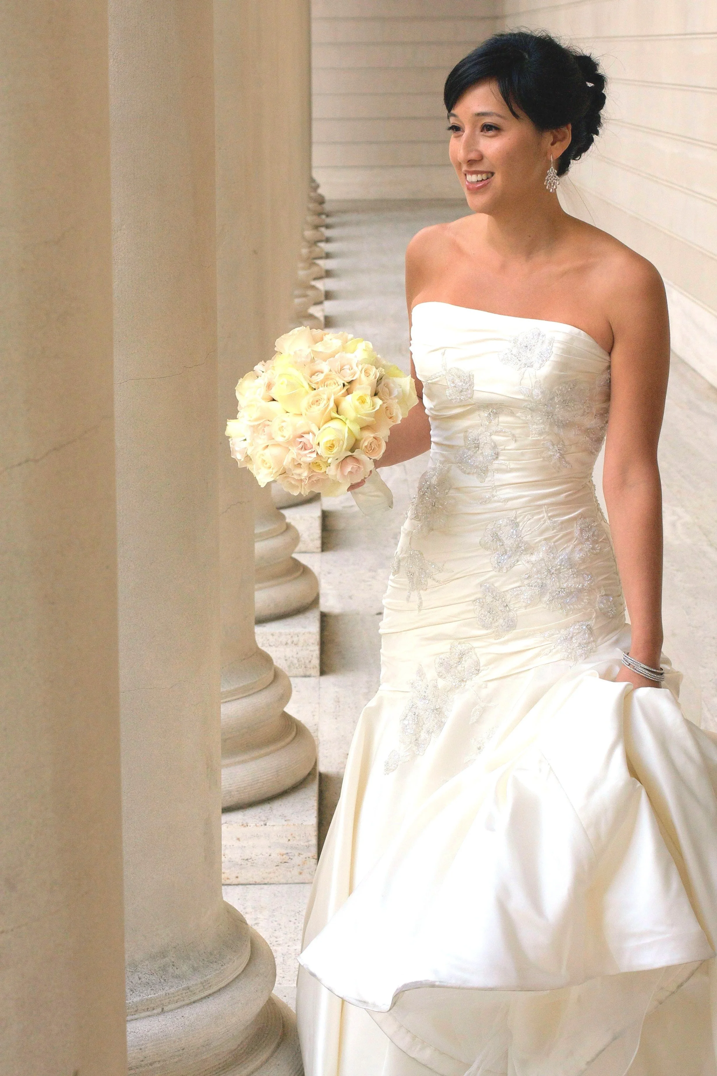 A smiling woman in a strapless white wedding gown holding a bouquet of light-colored roses and wearing sparkling earrings, standing outdoors beside columns with a wooden-paneled wall in the background.