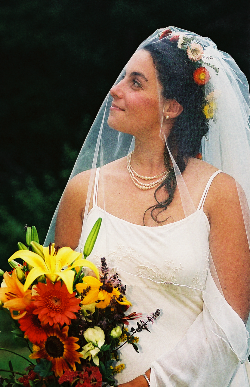 A smiling bride with dark hair wearing a white wedding dress, pearl necklace, and veil adorned with flowers, holding a colorful bouquet of yellow lilies, orange gerbera daisies, and other flowers, outdoors against a dark green background.