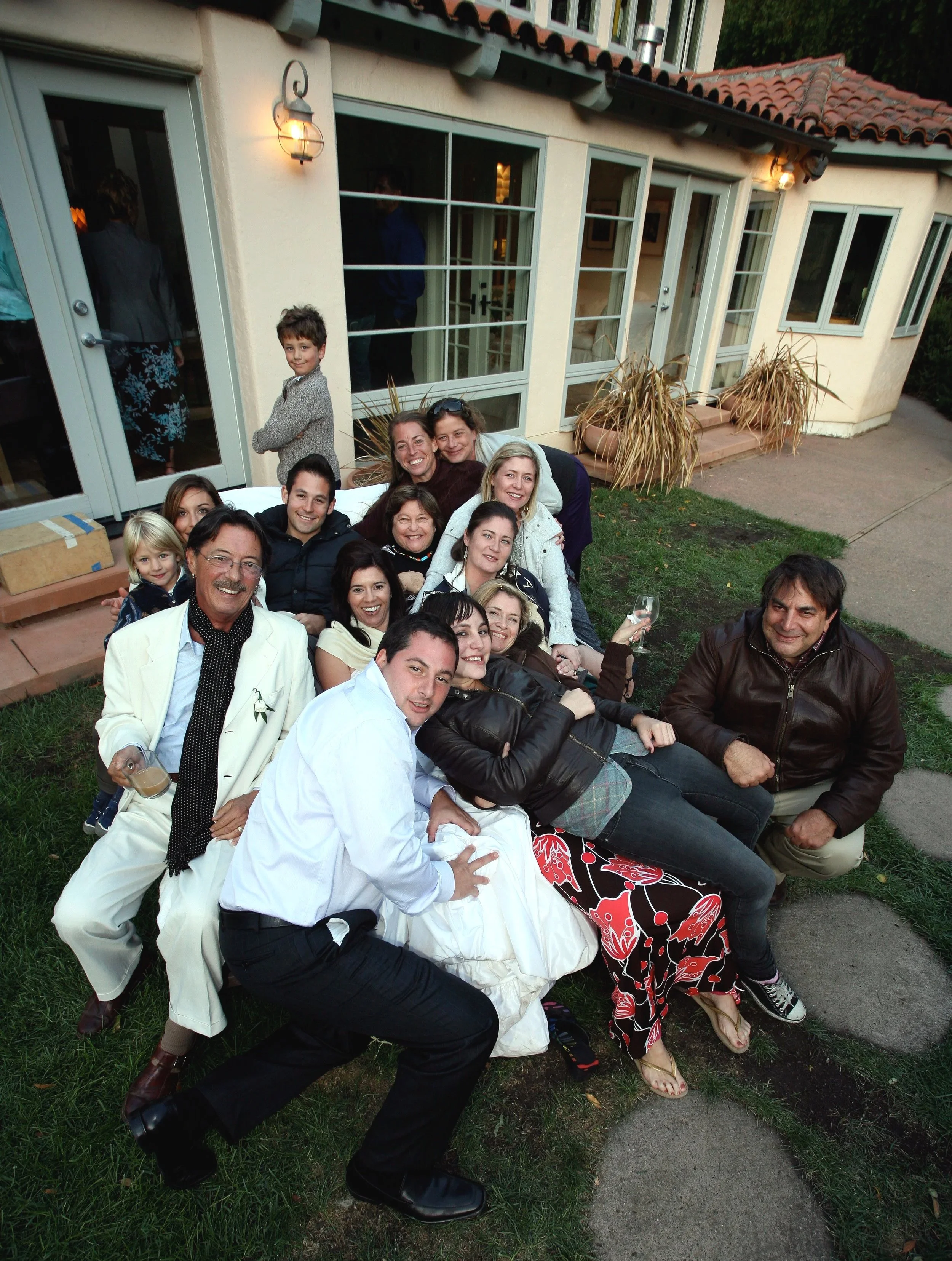 Newlyweds with a group of wedding guests together outside a house, with some sitting on the grass, smiling and enjoying the gathering during the evening.