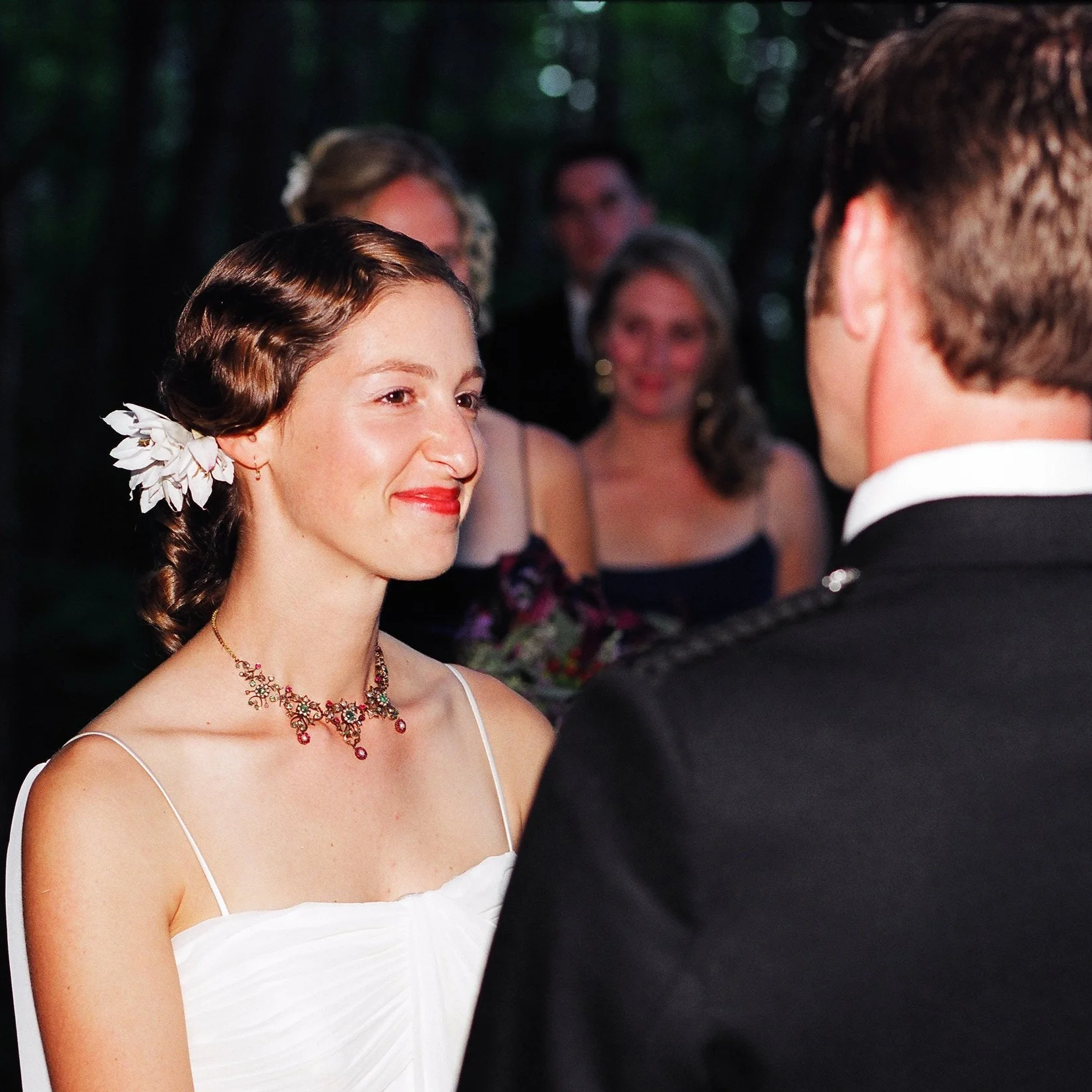 A woman with dark brown hair styled with a white flower hair accessory is wearing a white dress with thin straps and a red jewelry set, standing face-to-face with a man in a black tuxedo during an outdoor wedding ceremony. Four onlookers are blurred 