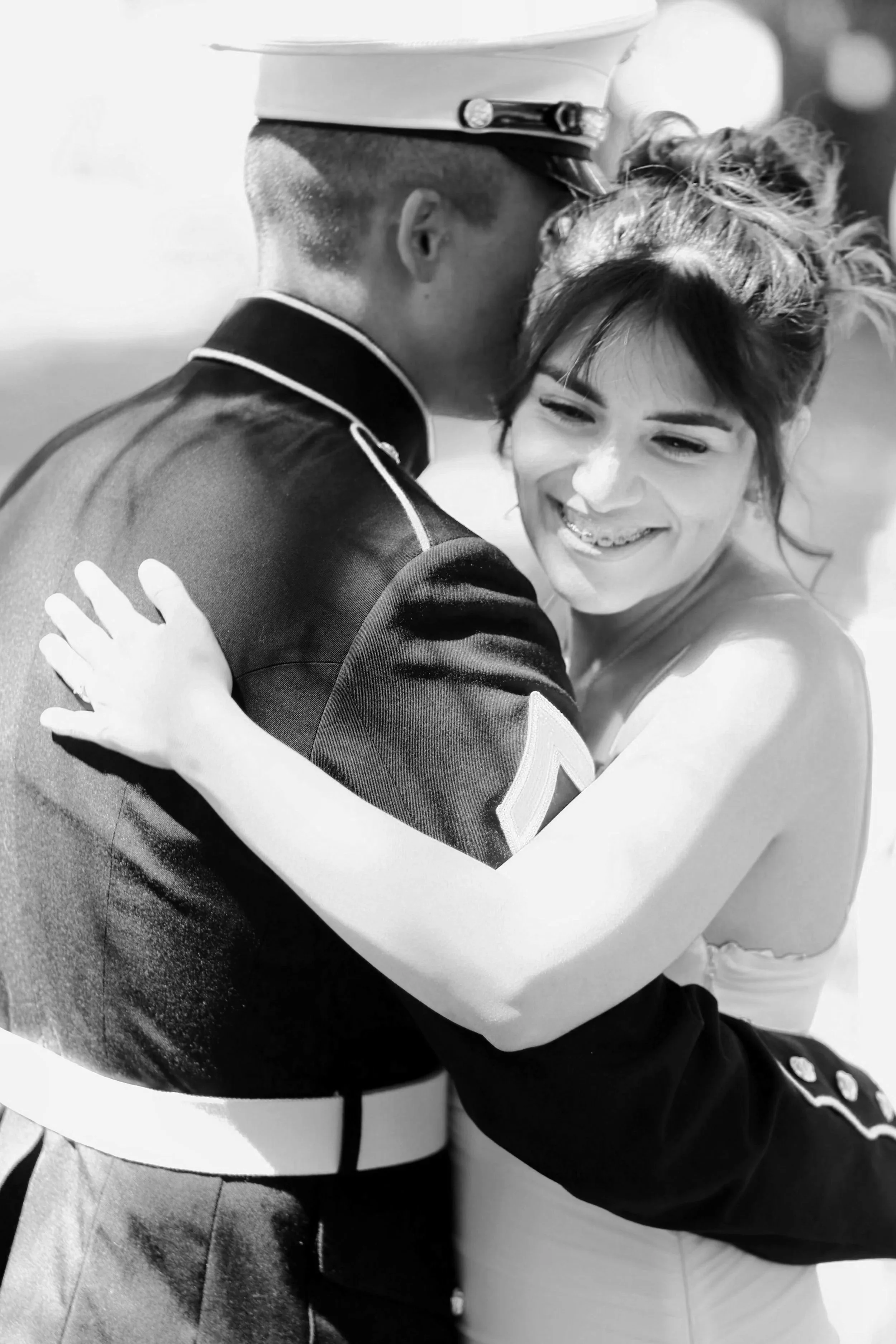 A black and white photo of a young newlywed couple hugging, the person on the left is in a uniform wearing a white hat and dark jacket, the person on the right has short hair with a braid and is smiling with braces.