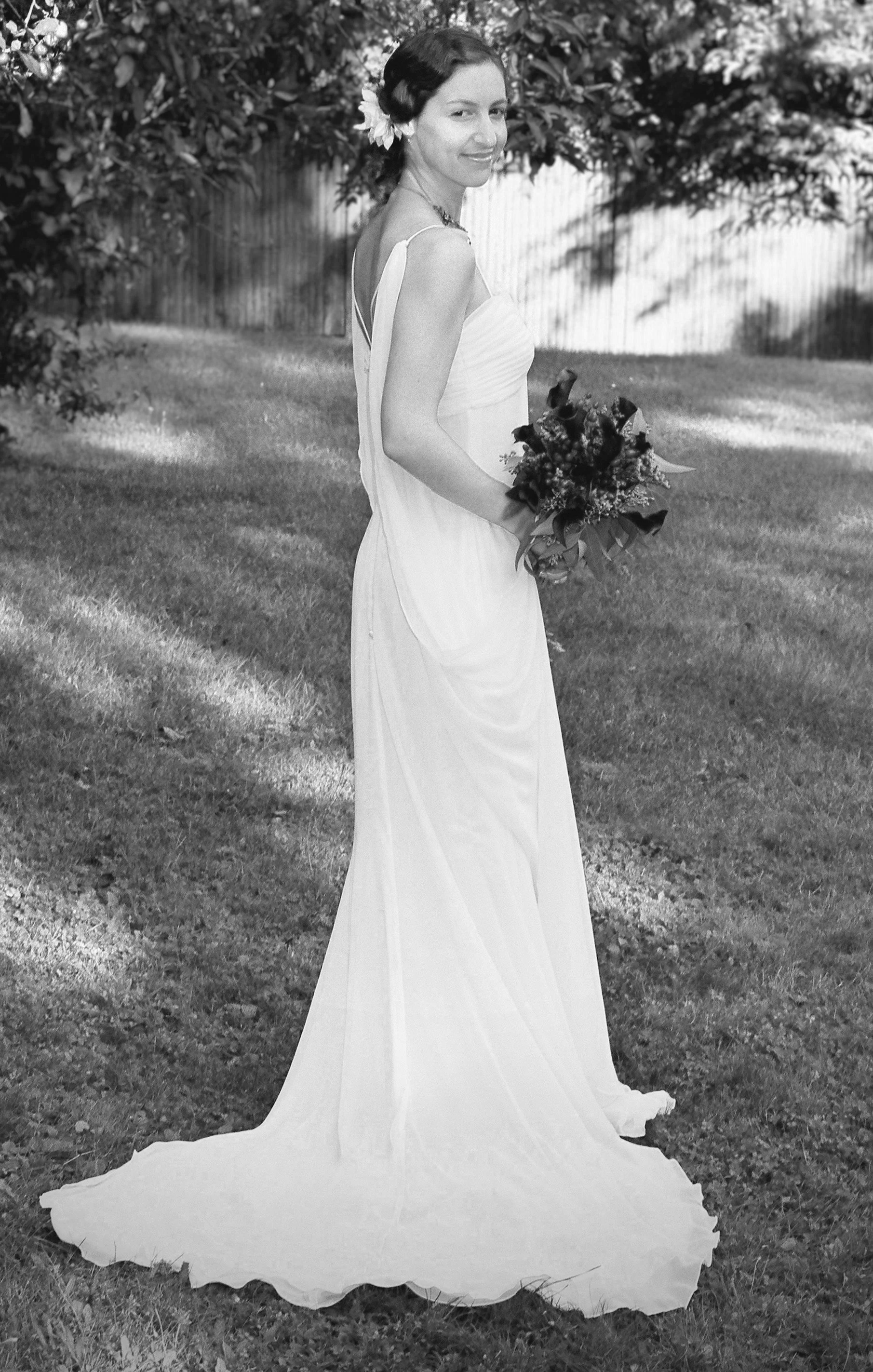 A black and white photo of a woman in a flowing dress holding a bouquet of flowers. She is standing outdoors on grass with trees and a wooden fence in the background, facing slightly to the right with a slight smile.