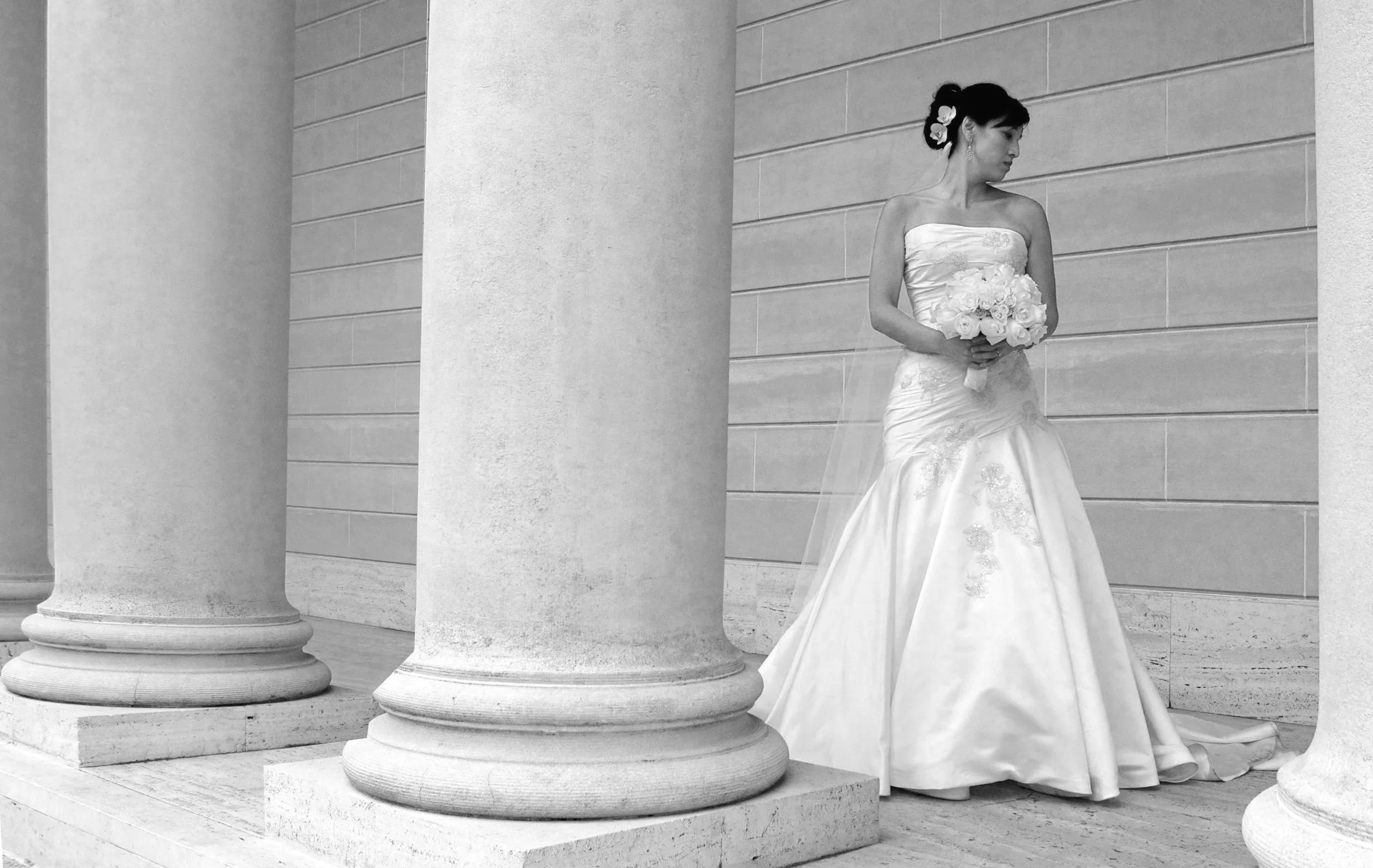 A woman in a wedding dress holding a bouquet of flowers stands between large stone columns against a textured stone wall.