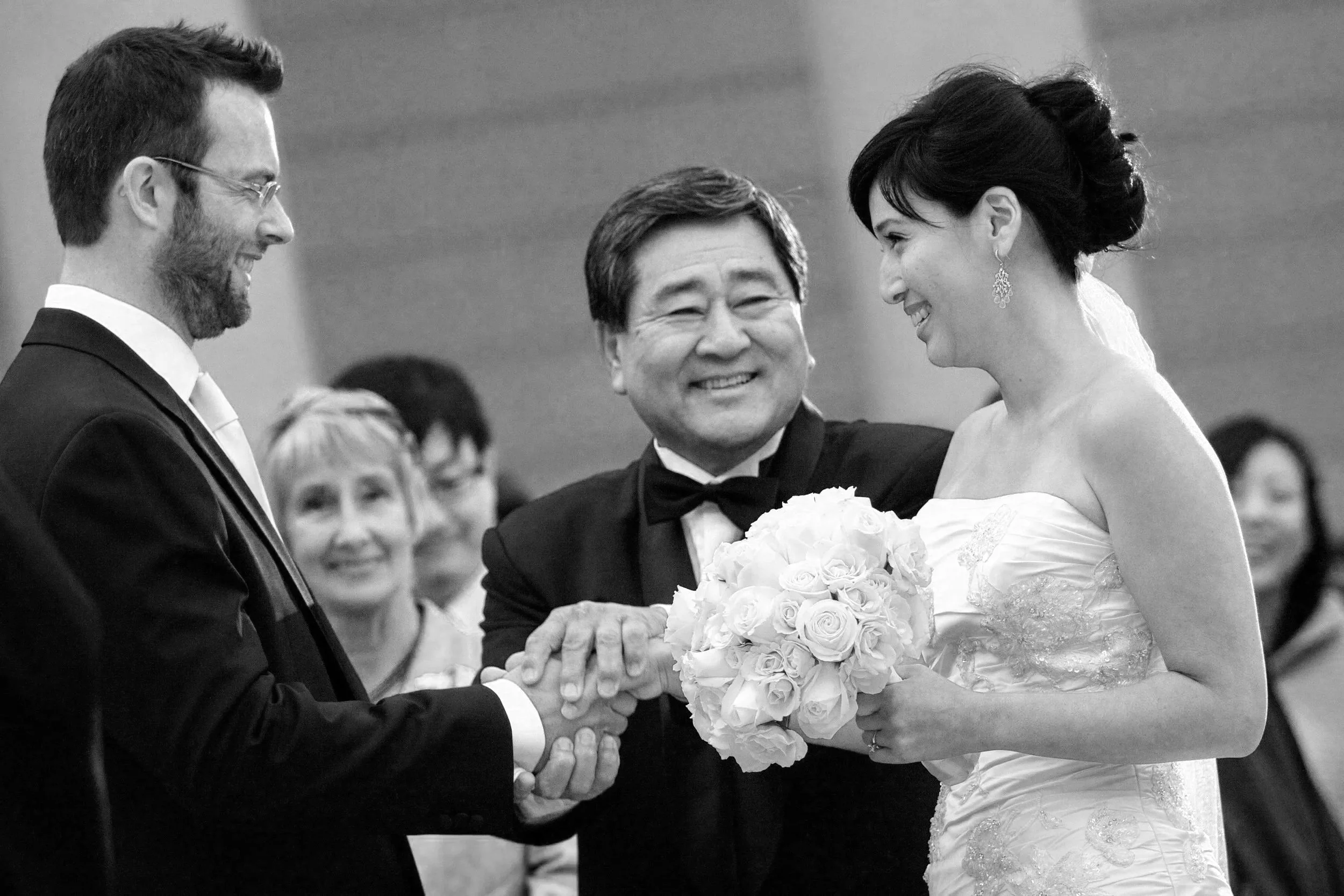 A black and white photo of a wedding ceremony with a bride and groom exchanging rings, surrounded by a smiling officiant and guests.