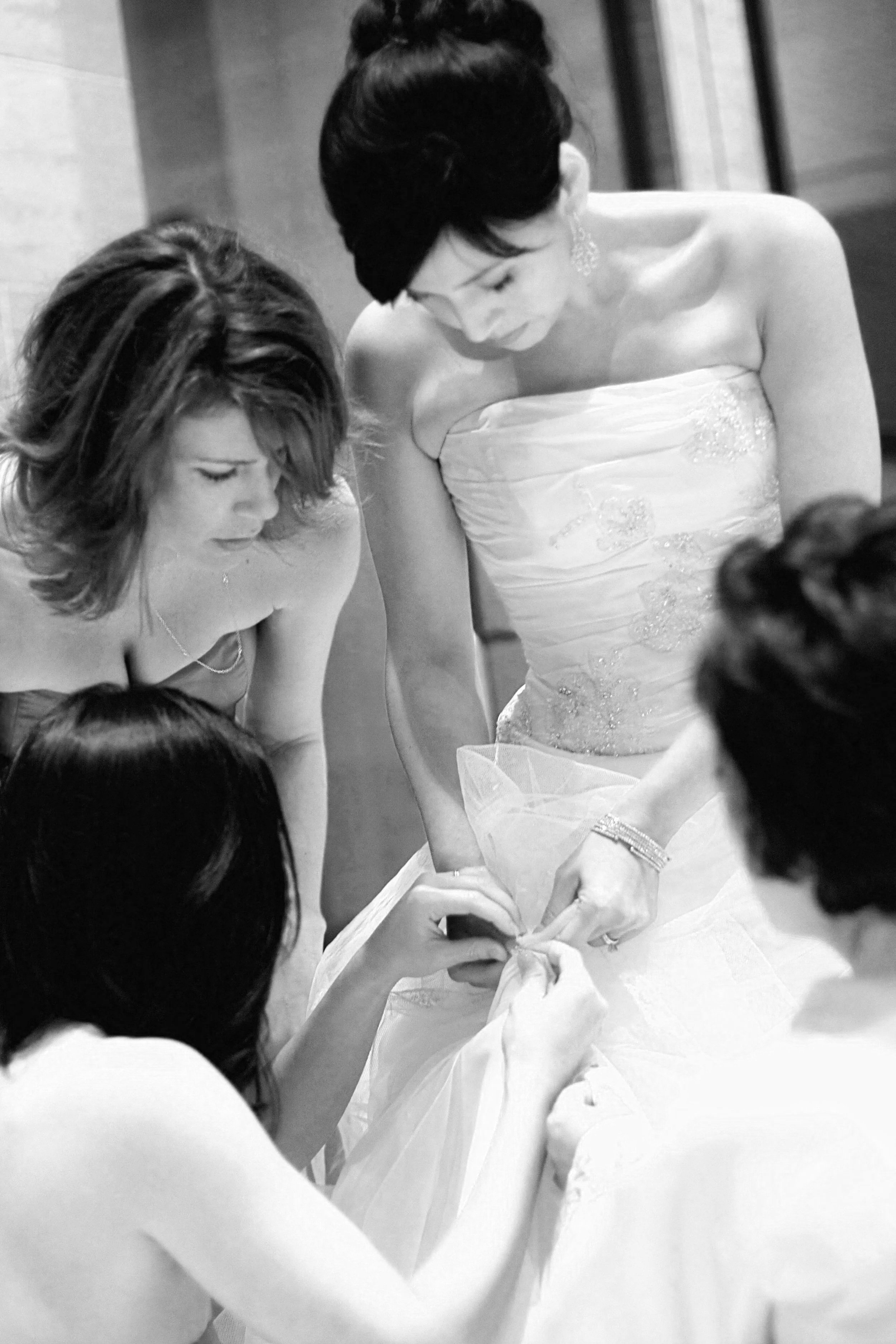 Women in elegant dresses assisting with a wedding dress, likely at a wedding or bridal boutique.
