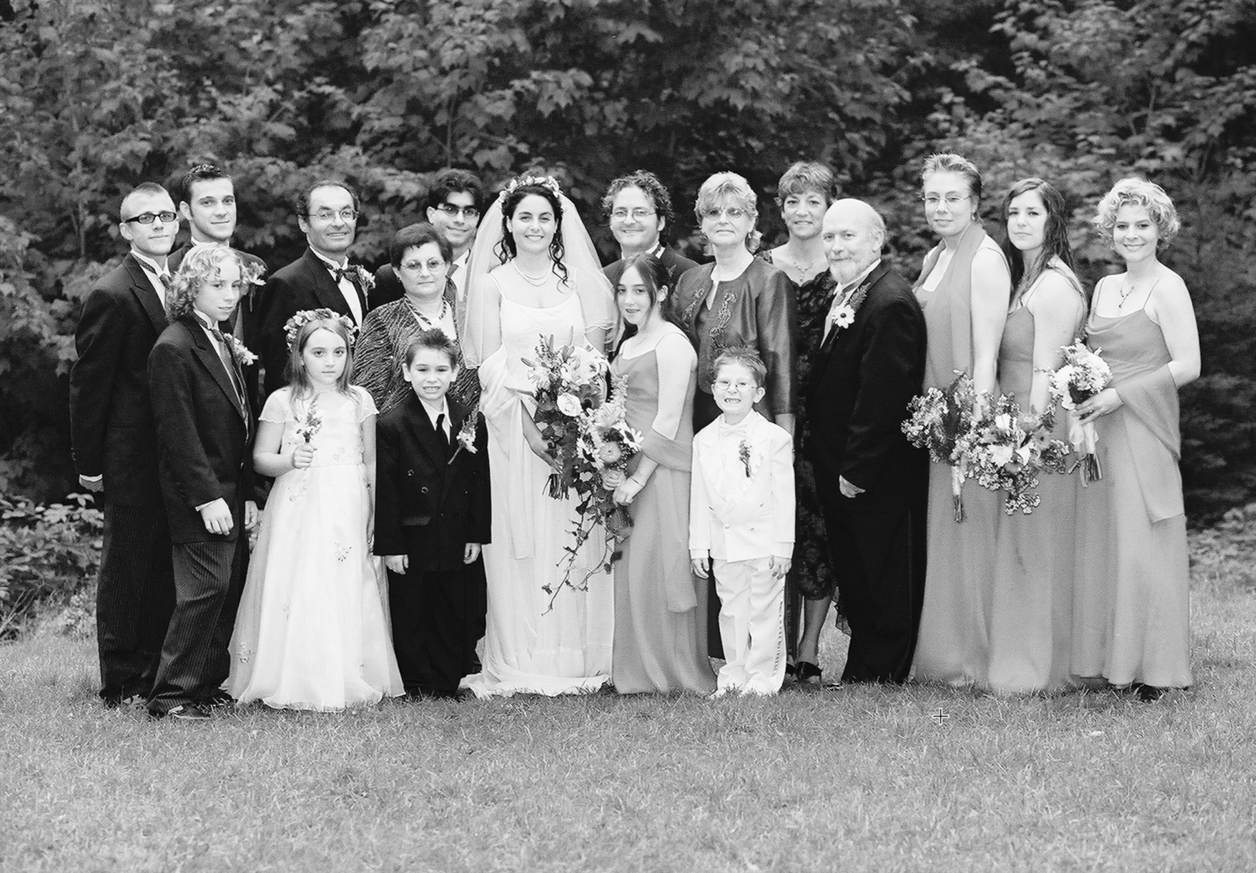Black and white photo of a wedding party outdoors with trees in the background, including a bride, groom, children, and adults, all dressed in formal attire, standing on grass.