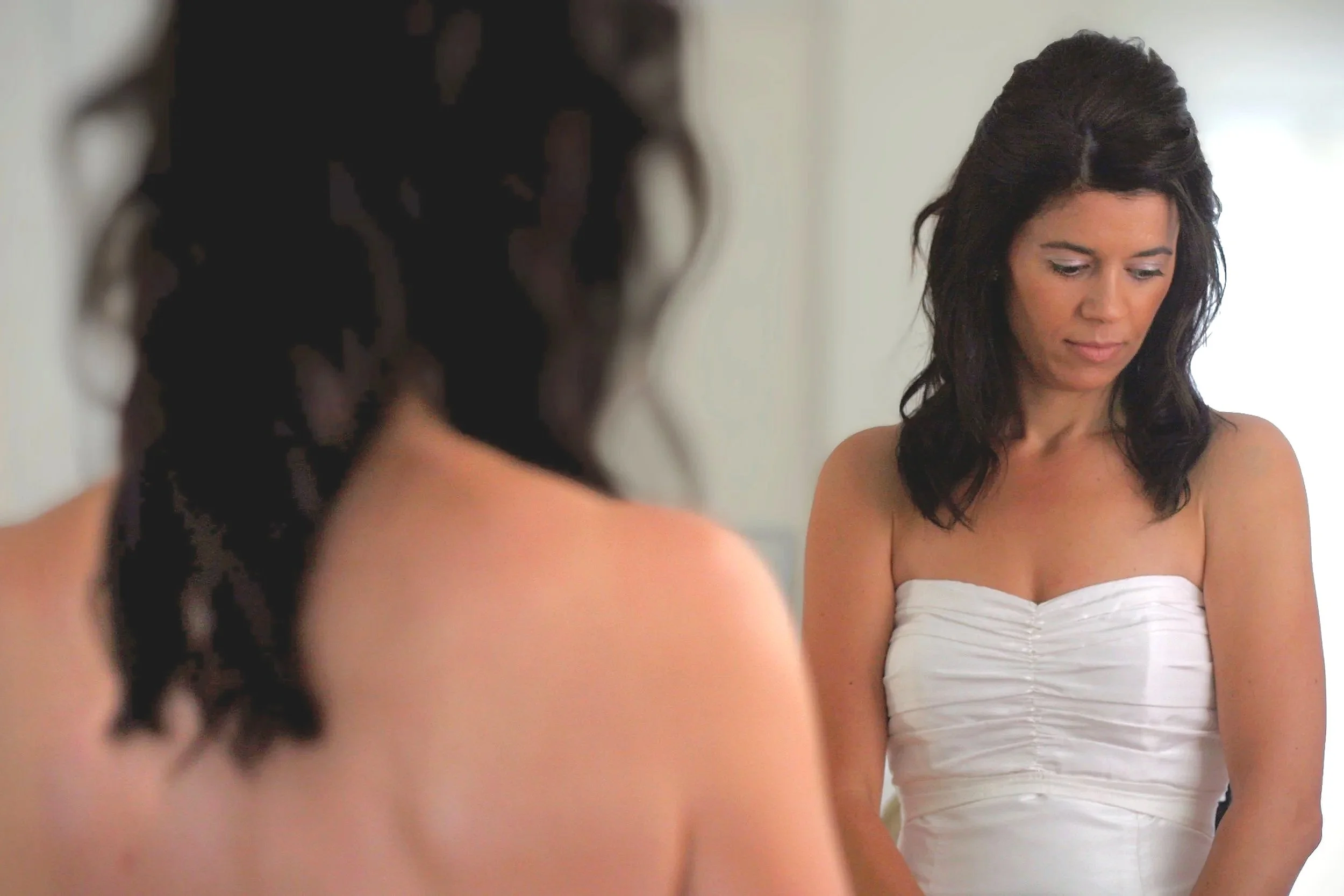 A woman with dark brown wavy hair wearing a white strapless dress looking down, reflected in a mirror.