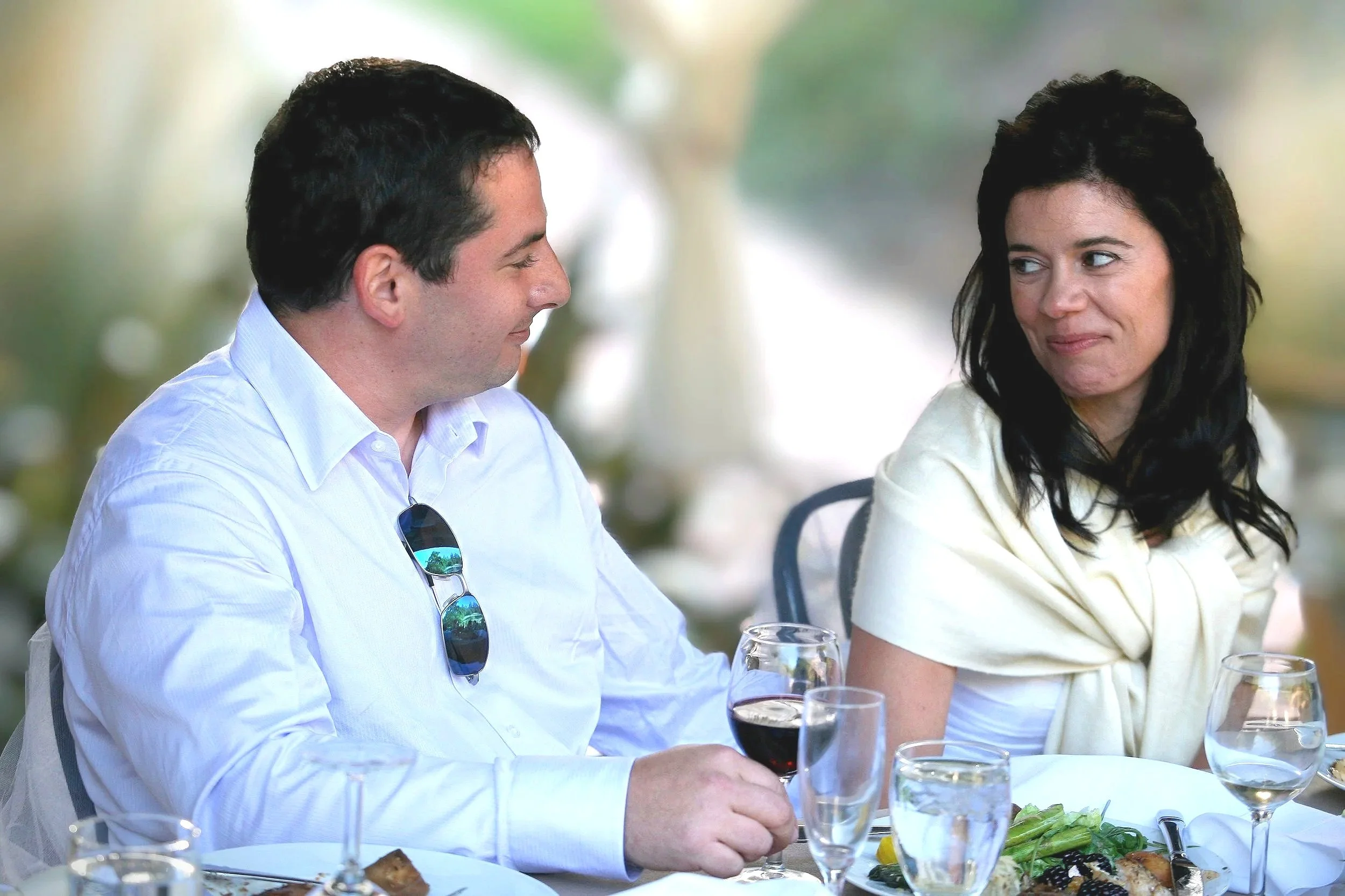 A man and woman sit at a table outdoors during daytime, engaged in conversation. The man is wearing a white shirt with sunglasses hanging from his collar, and the woman has dark, wavy hair and is wearing a white top. There are glasses of wine and pla