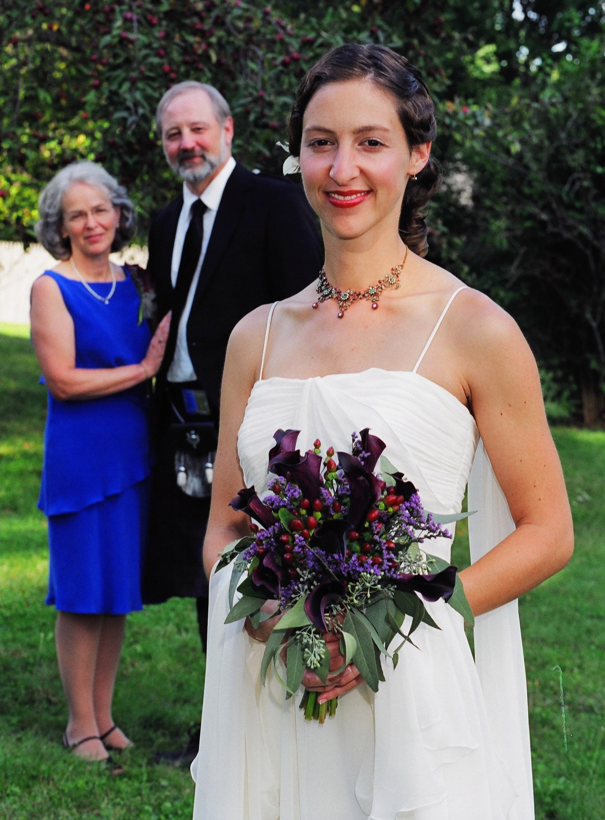 A young woman in a white dress holding a bouquet of purple and red flowers, standing outdoors with green trees in the background. Two older adult parents, a woman in a blue dress and a man in a black suit, are blurred in the background.
