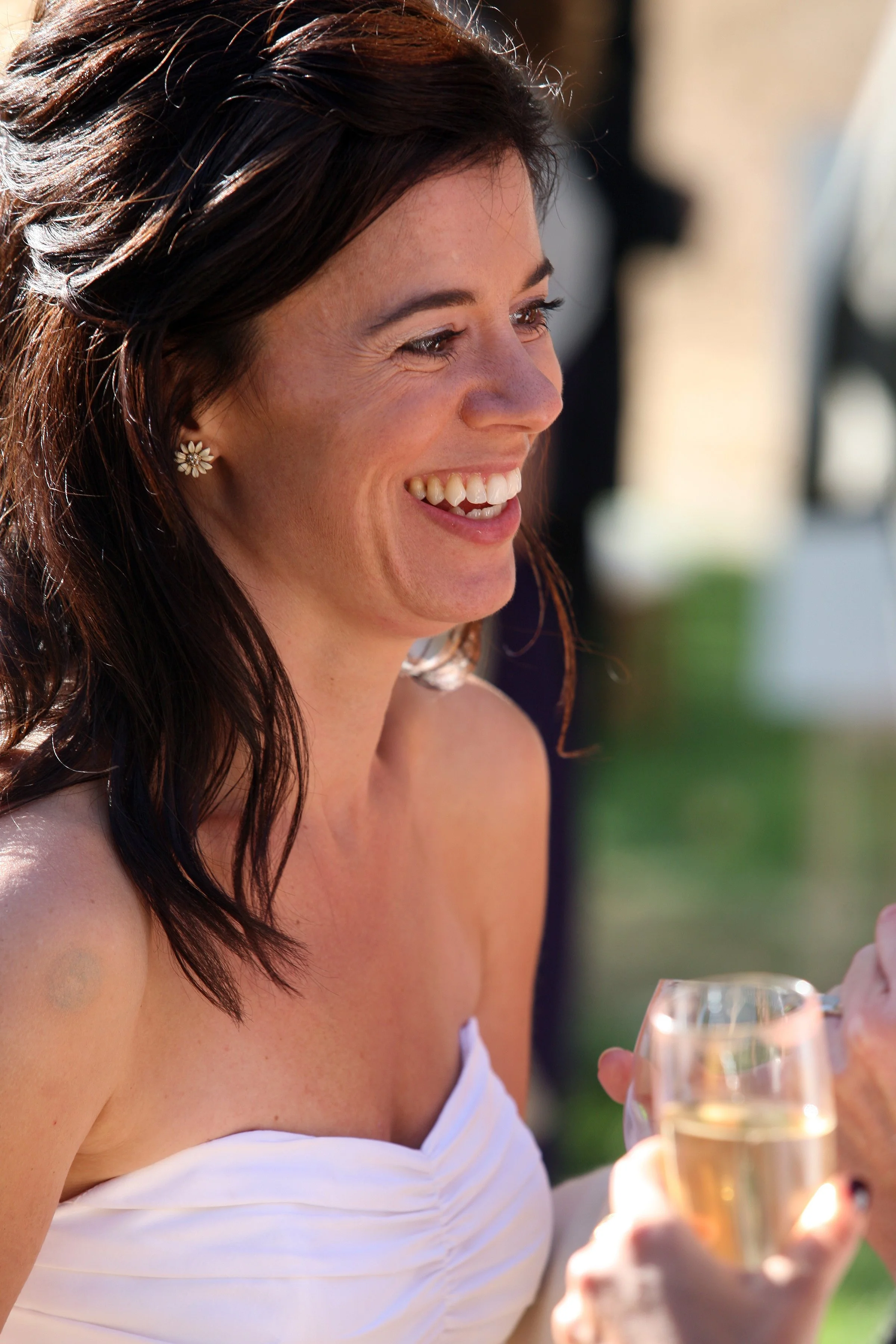 A woman with dark brown hair, wearing floral earrings, smiling and holding a glass of white wine at an outdoor event.