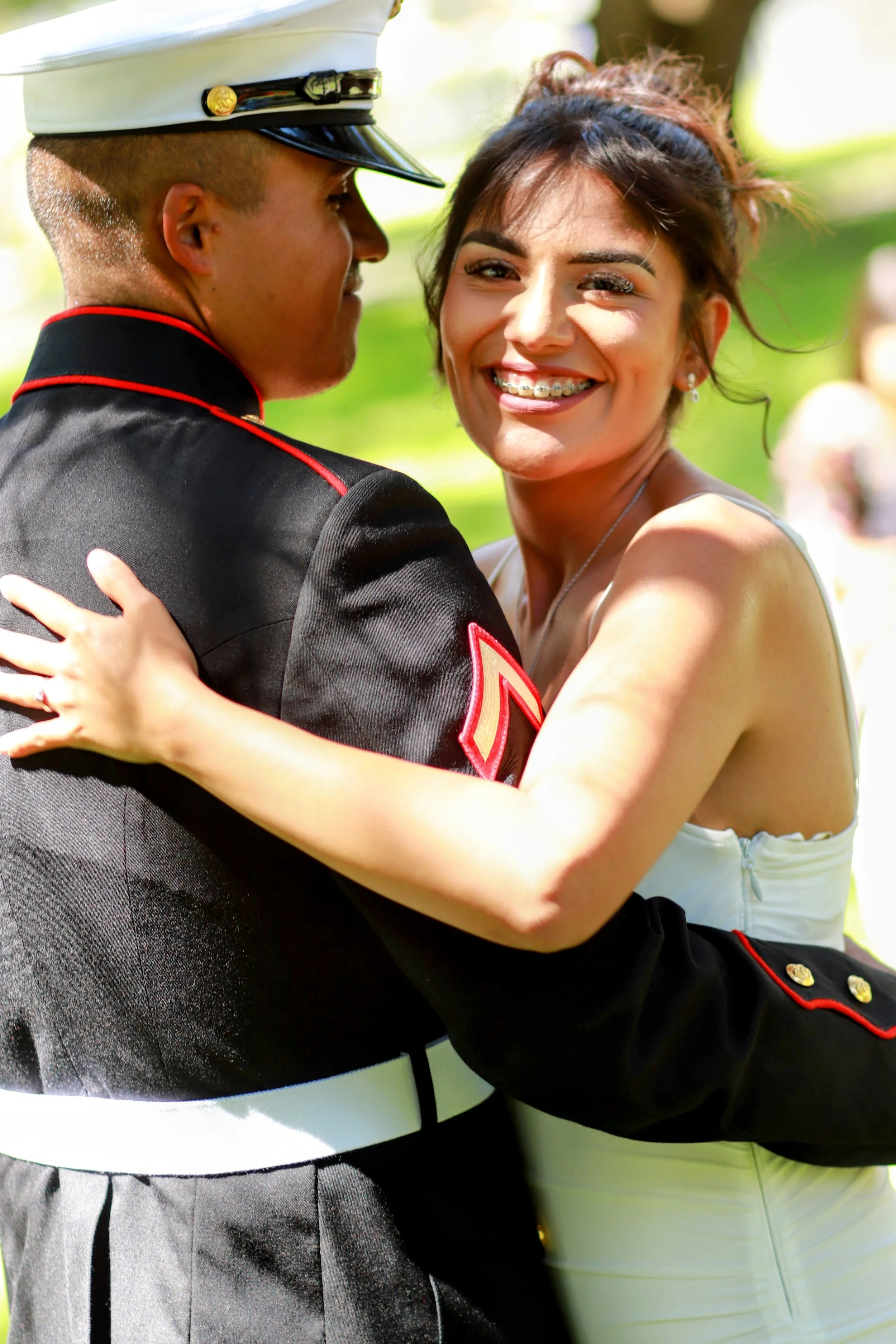 Newlyweds embracing outdoors, the man in a military uniform with a white hat and the woman smiling with braces, her hair tied up, wearing a white dress.
