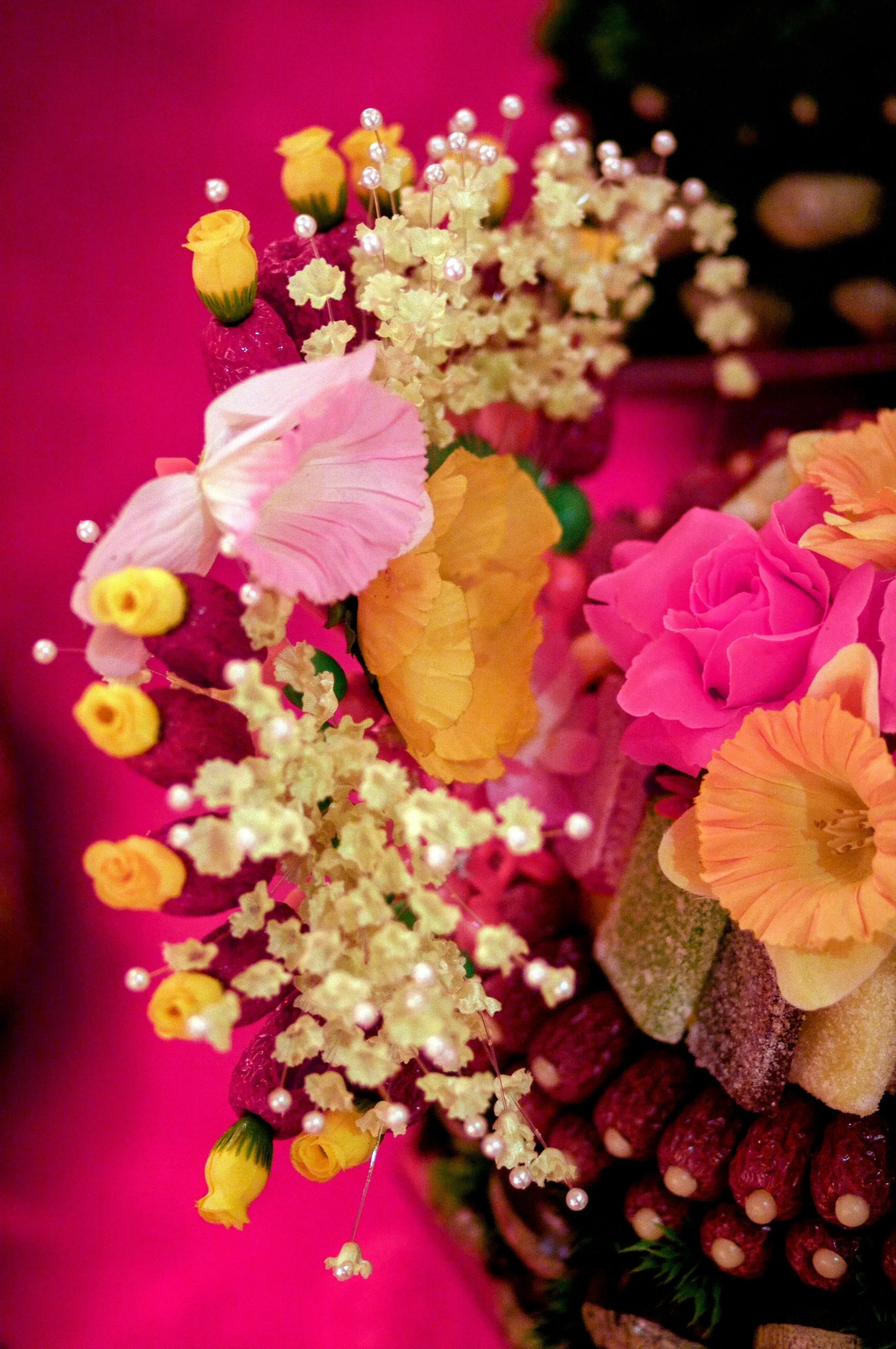 Close-up of a floral arrangement with pink, yellow, and white flowers, pearl accents, and greenery against a pink background.