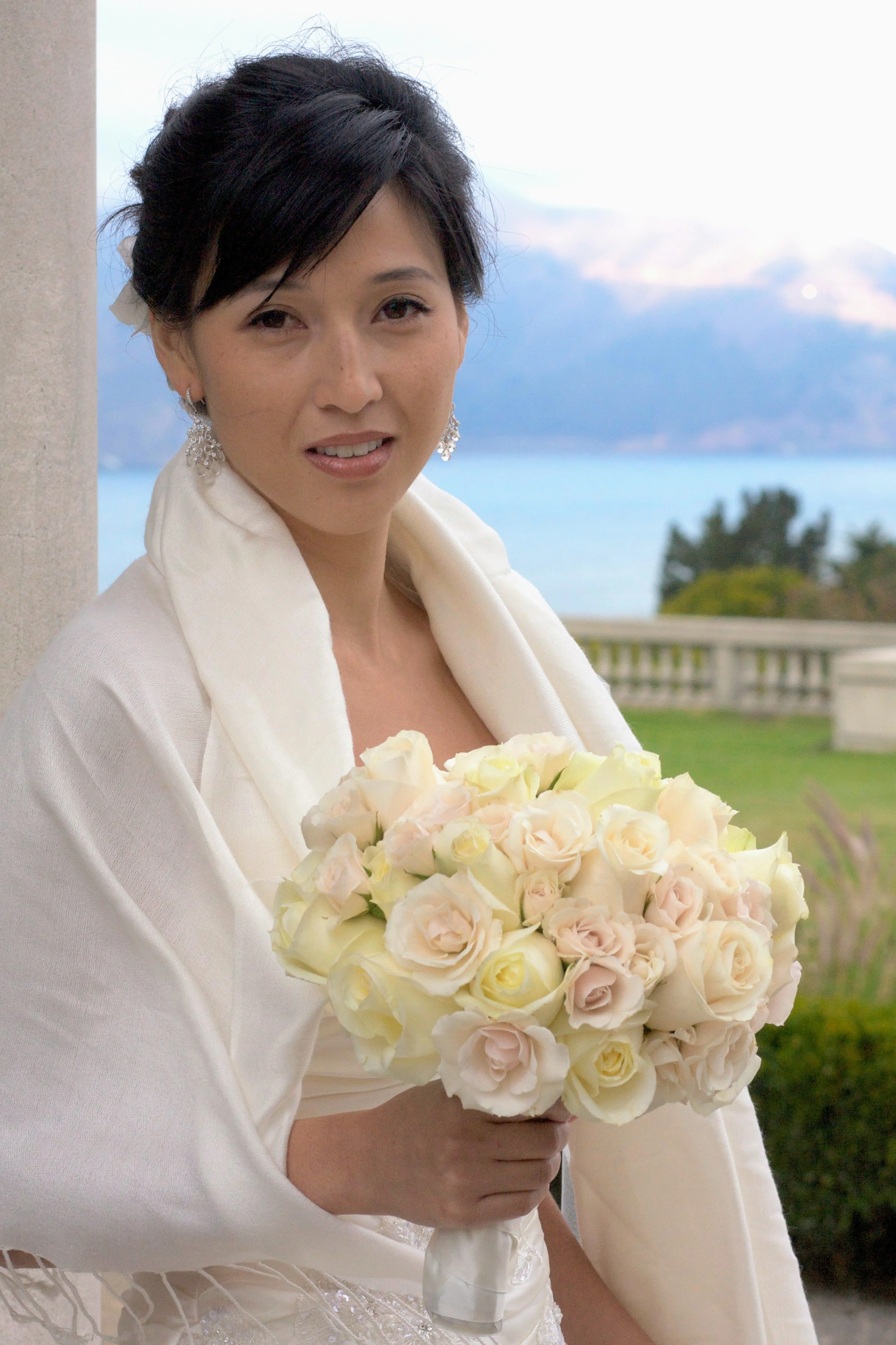 A woman dressed in white holding a bouquet of pale pink and white roses outdoors near a stone column, with a waterfront and cloudy sky in the background.