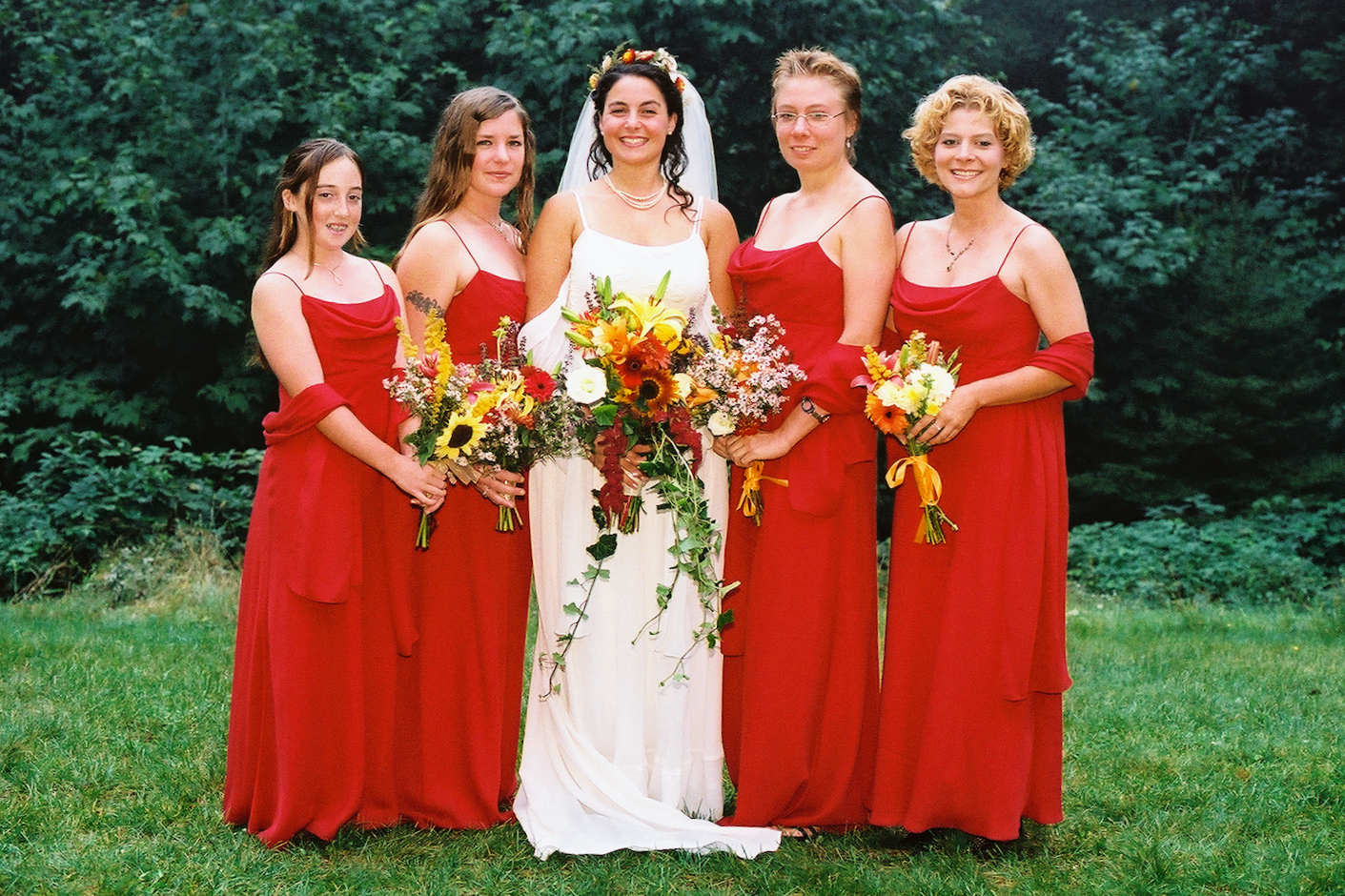 A group of six women dressed in formal dresses, standing outdoors on grass with trees in the background. The woman in the center is wearing a white wedding gown with a floral crown and holding a bouquet. The other five women are wearing red dresses a