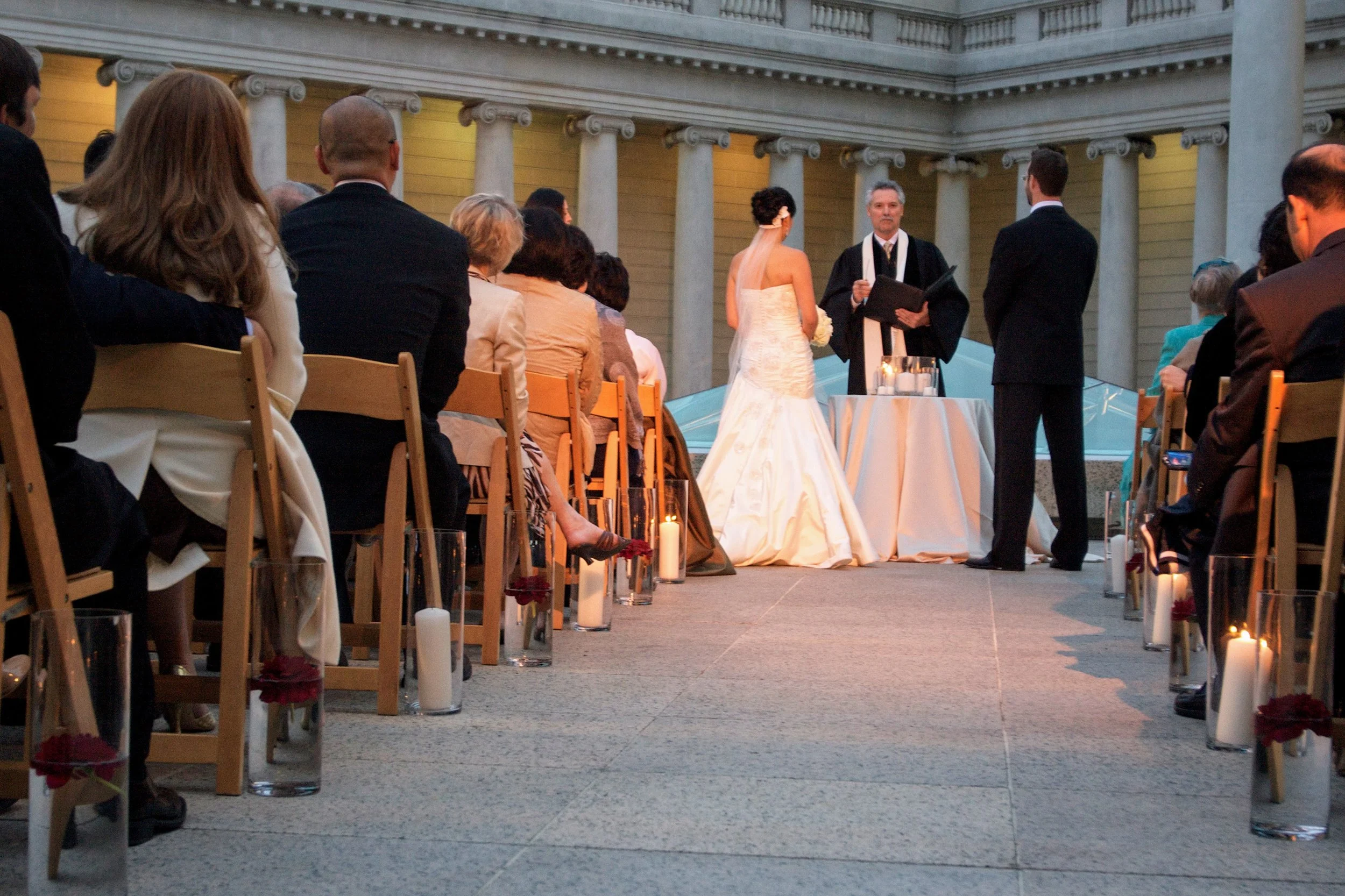 A wedding ceremony taking place outdoors, with a bride and groom standing in front of a clergyperson, surrounded by seated guests, candles, and floral decorations.