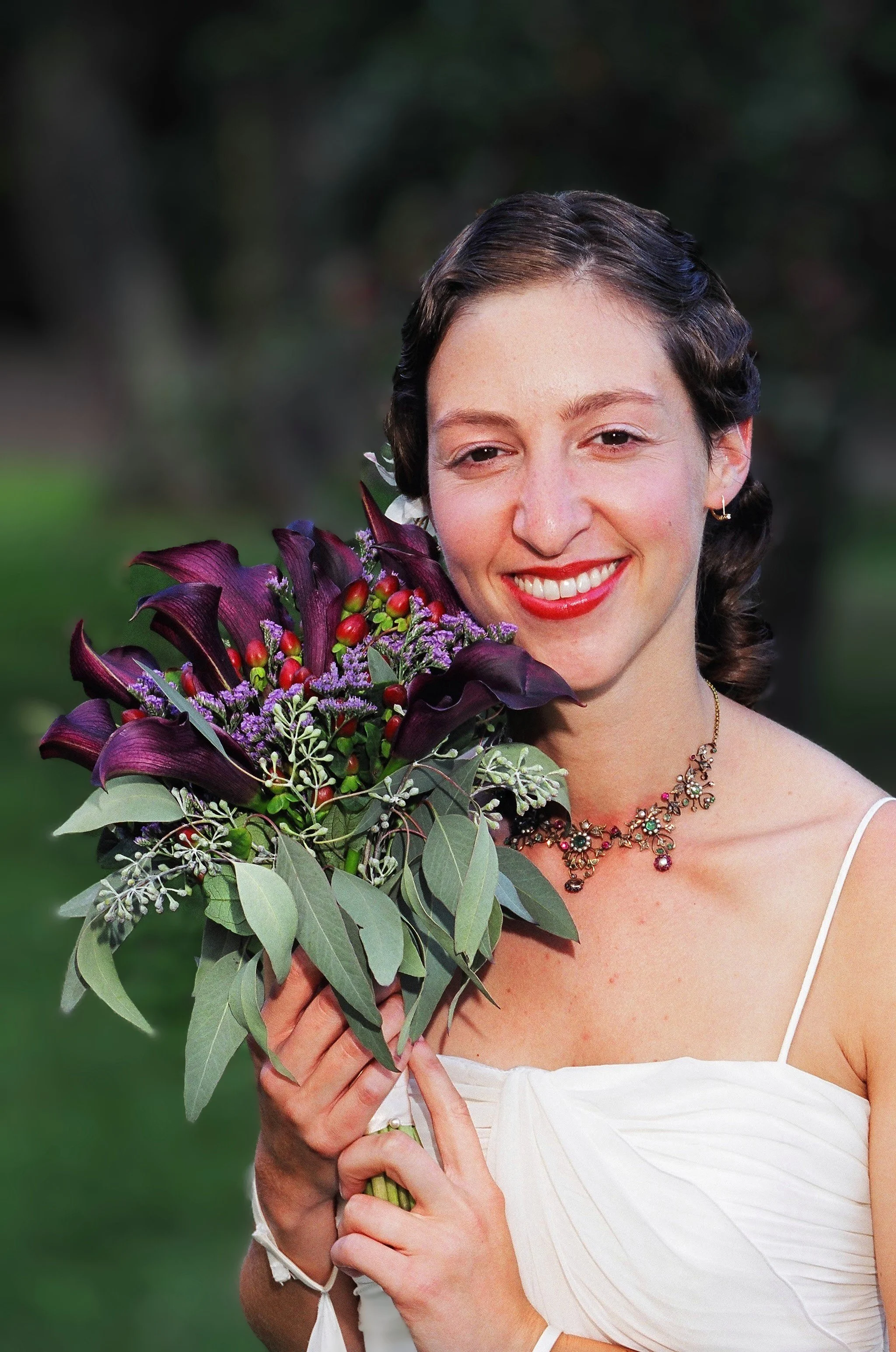 A woman in a white dress holding a bouquet of purple and red flowers with green leaves, outdoors with a blurred natural background, smiling.