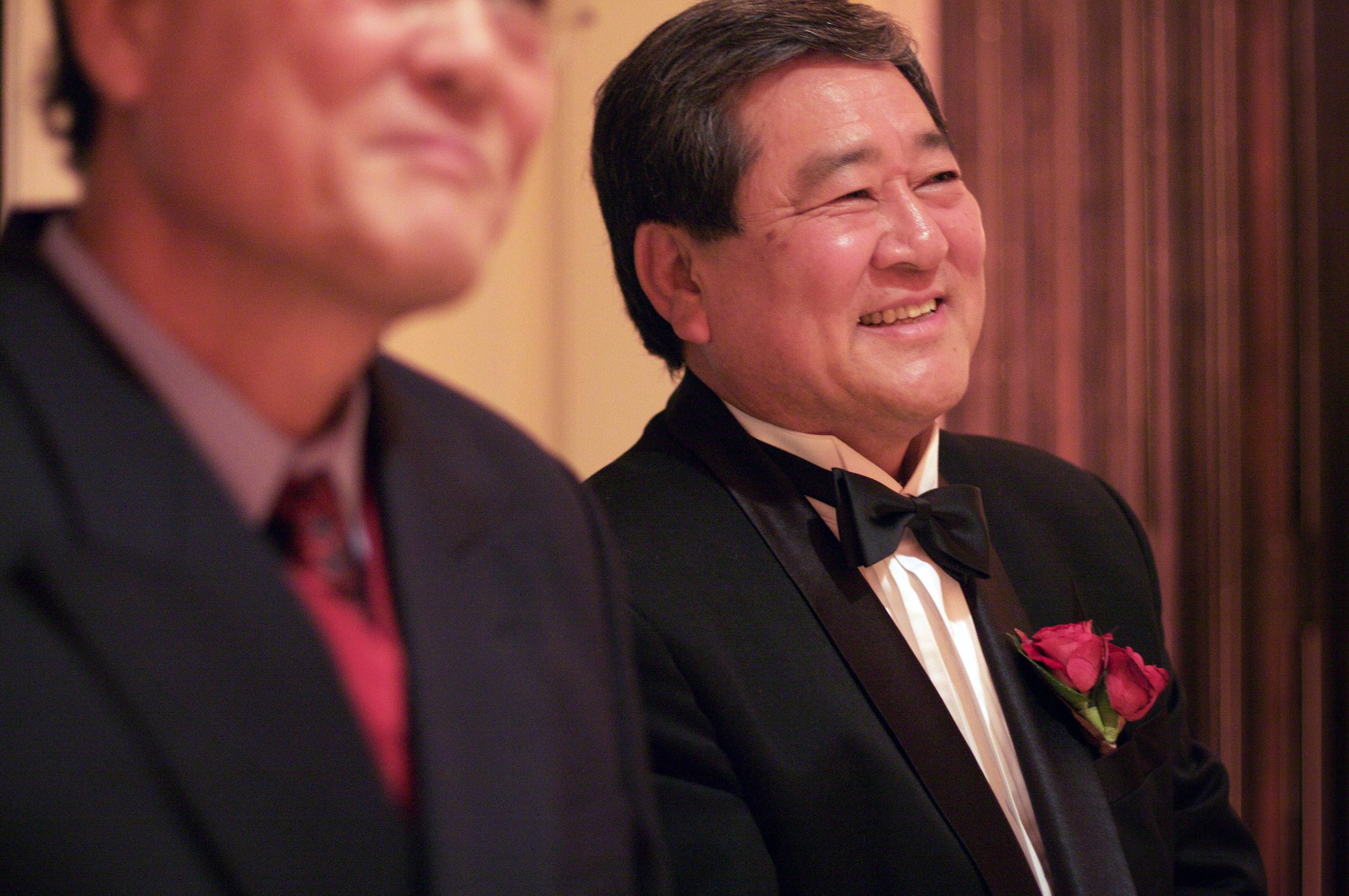 Two men in tuxedos, one with a red flower boutonniere, smiling during a formal event. The man on the right is in focus, while the other is blurred.