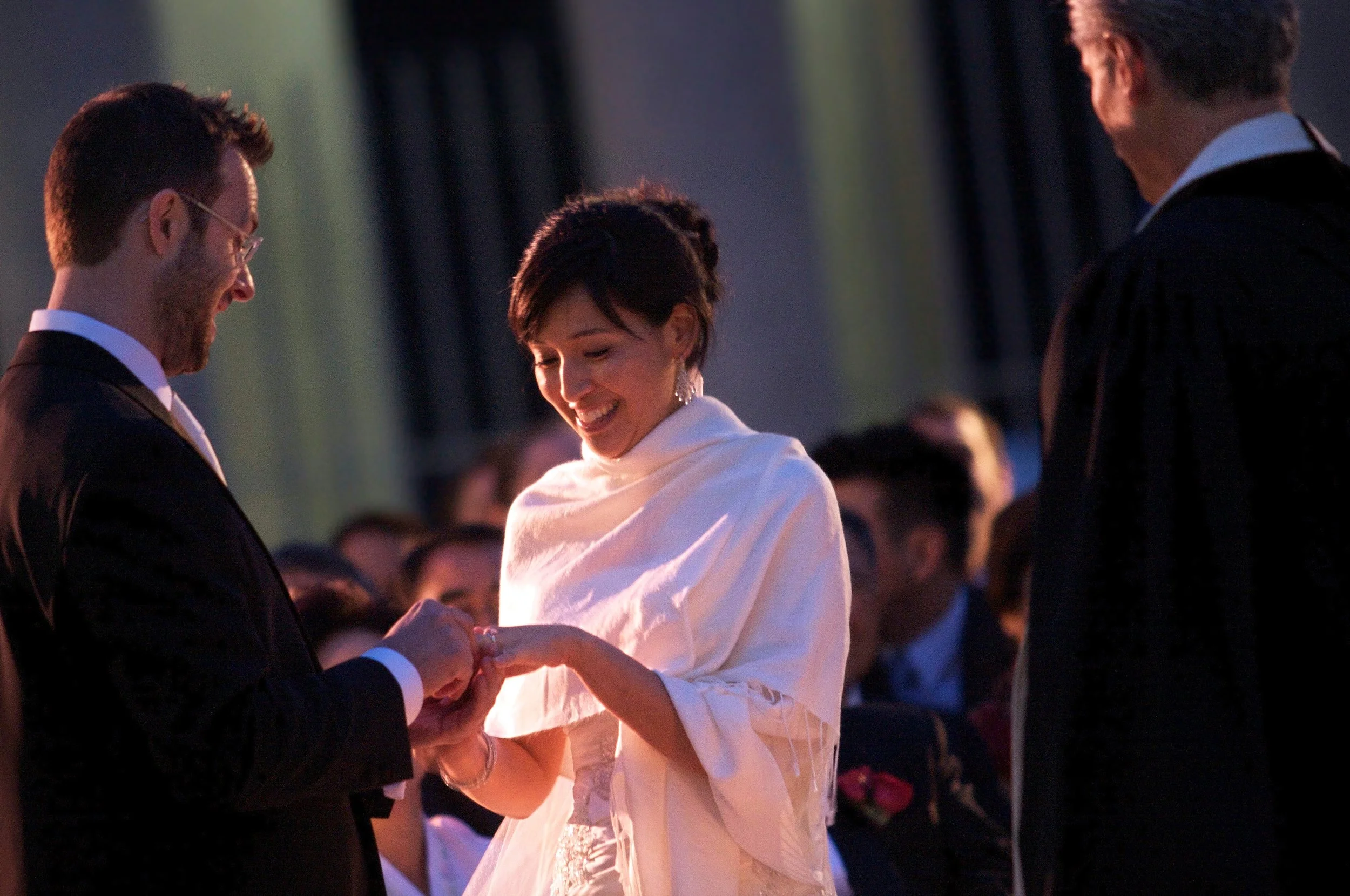 A couple is exchanging wedding vows during a ceremony, with a woman in a white dress and a man in a black suit, in a dimly lit indoor setting with guests in the background.