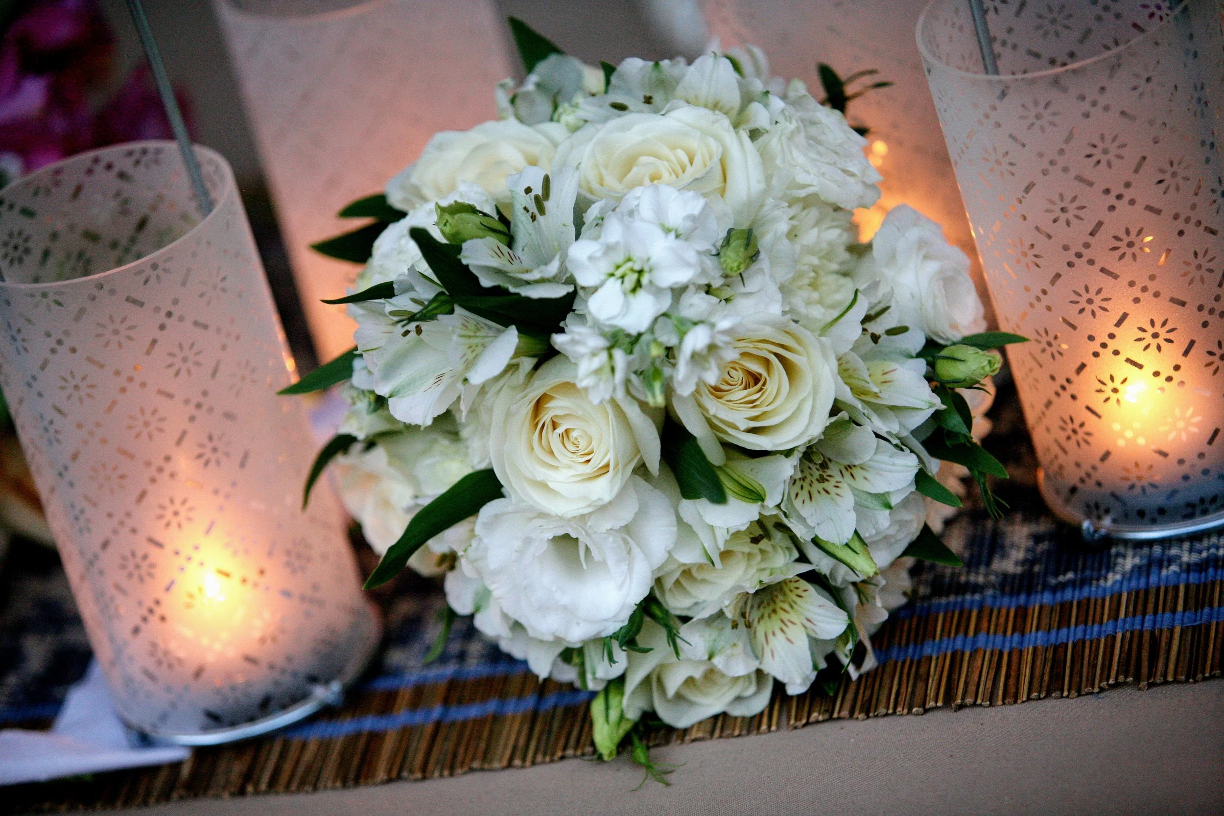 A bouquet of white roses and white lilies surrounded by green leaves, placed on a woven mat, flanked by two white perforated candle holders with lit candles inside.