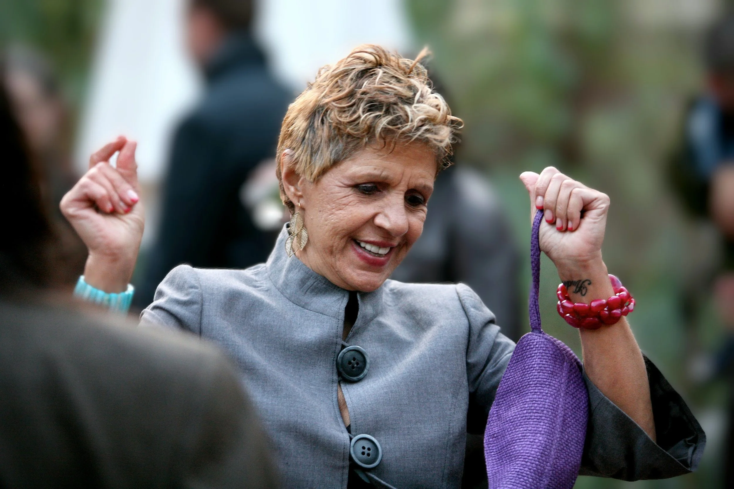 A woman with short blonde hair and large earrings smiles as she holds her arms up in a dancing pose, wearing a gray jacket with large buttons. She is holding a purple cloth and wearing red jewelry. The background is blurred with other people and natu