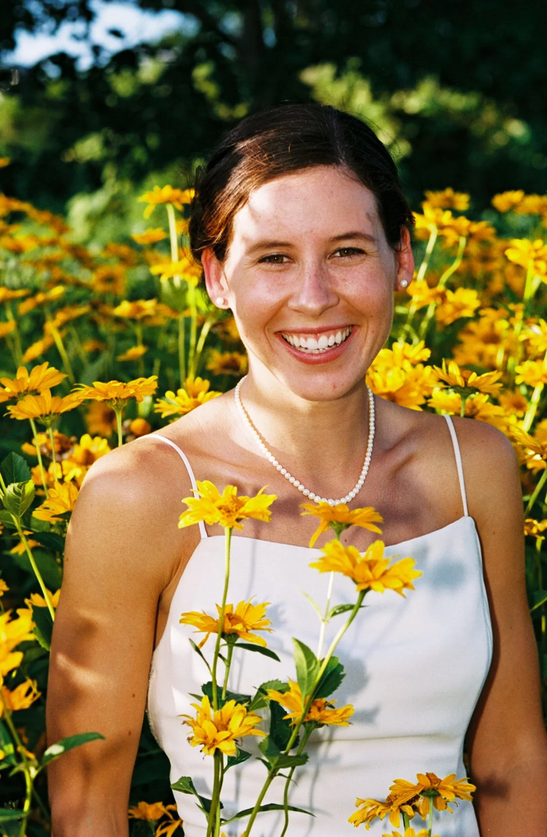 A woman with short brown hair smiling in a field of yellow flowers, wearing a white dress, pearl earrings, and a pearl necklace.