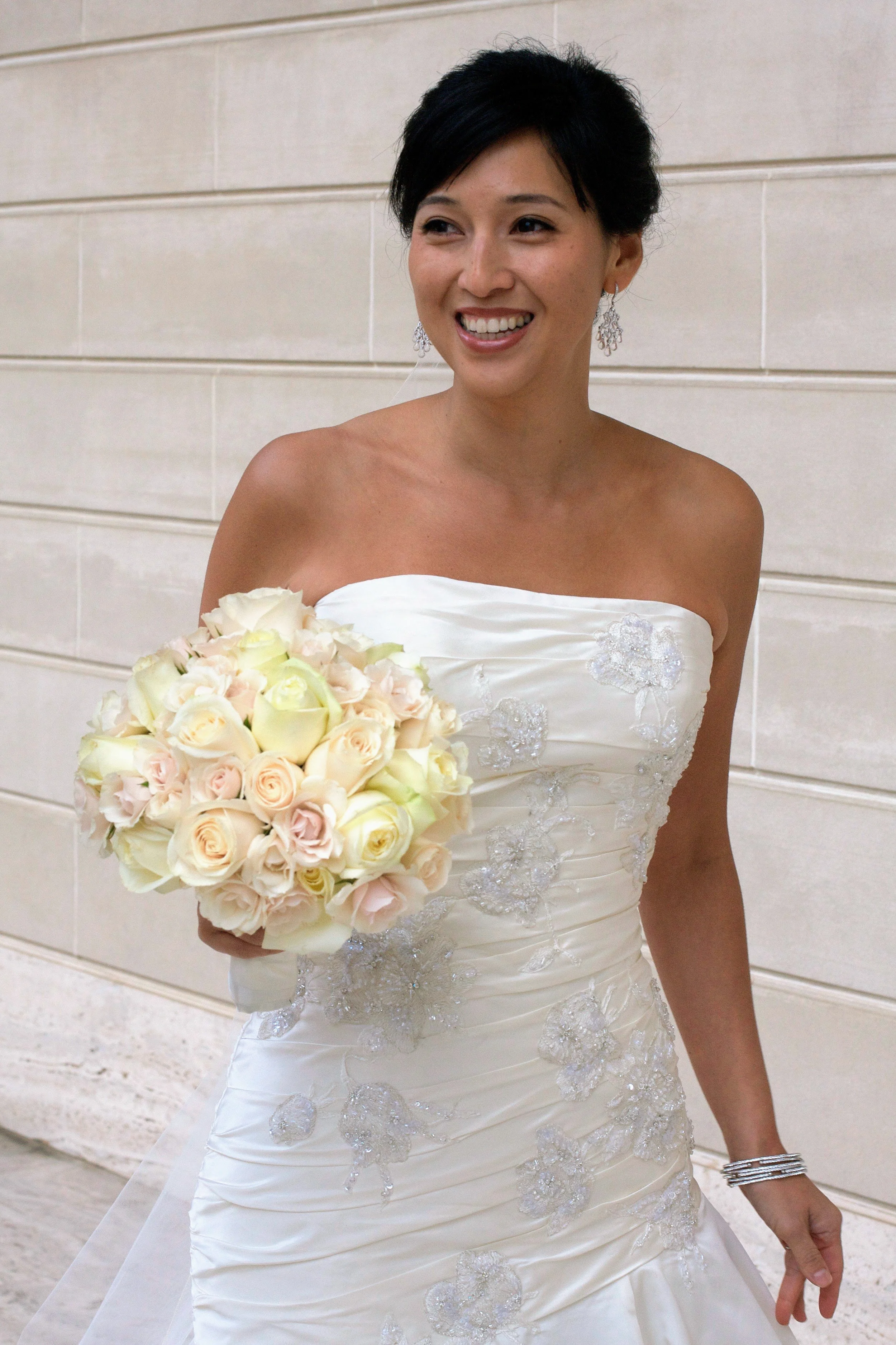 A woman in a white wedding dress holding a bouquet of white and pink roses, smiling, standing against a beige tiled wall.