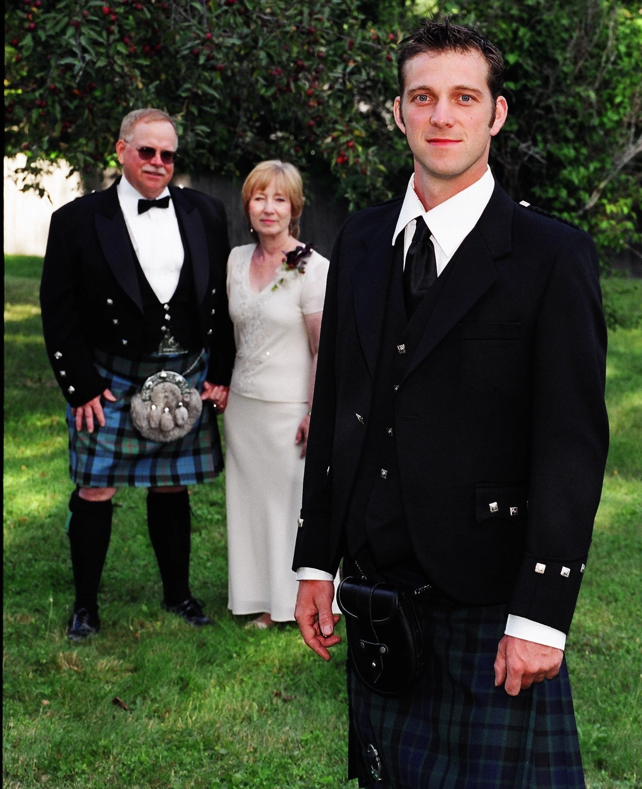 A group of four people dressed in formal attire standing outdoors on grass with trees in the background. One man in the foreground wearing a black tuxedo and tartan kilt, holding a black bag. A young man and an older couple standing behind him, with 
