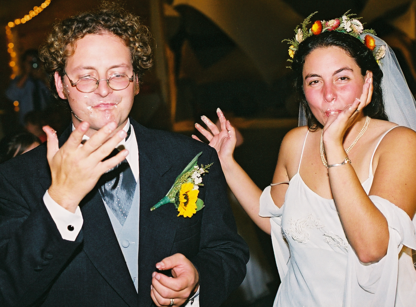 A man with glasses and a woman in a wedding dress are at a celebration. Both are covered in cake and have joyful expressions. The man is wearing a tuxedo with a sunflower boutonniere, and the woman is wearing a floral crown and a pearl necklace.