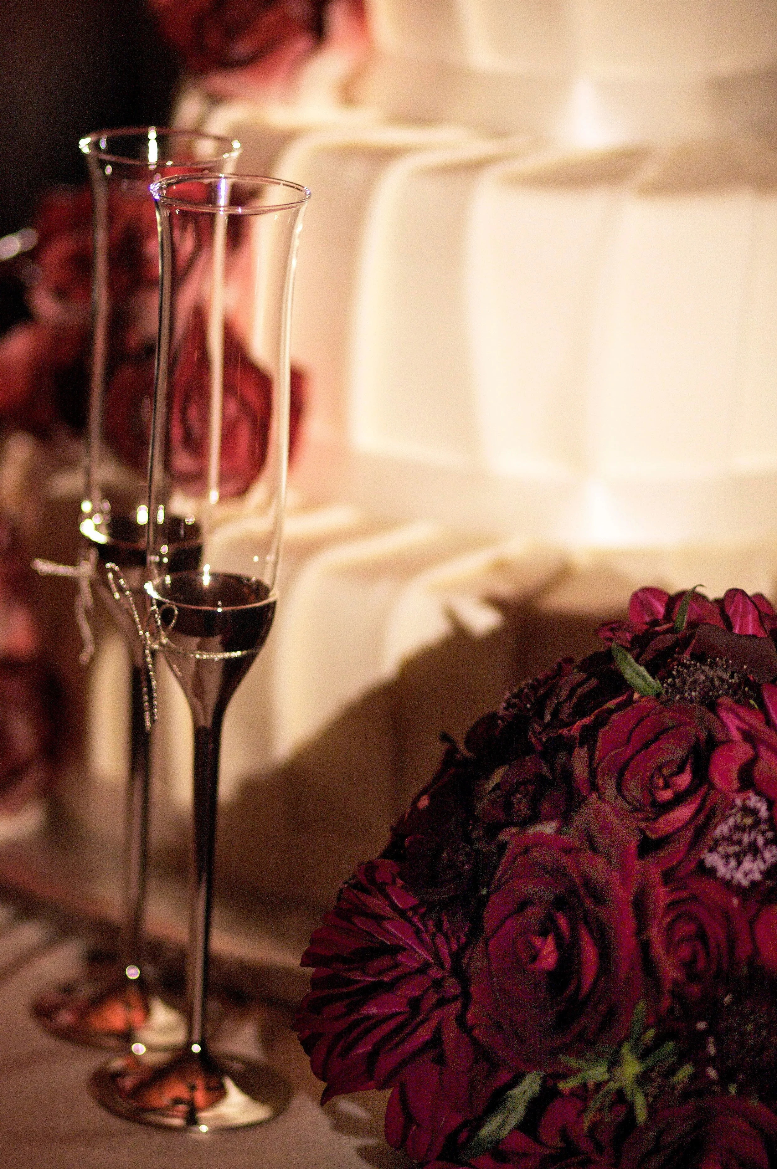 A close-up of two tall, empty champagne flutes with a glass bowl of purple and pink flowers in the foreground, and a blurred background with soft lighting.