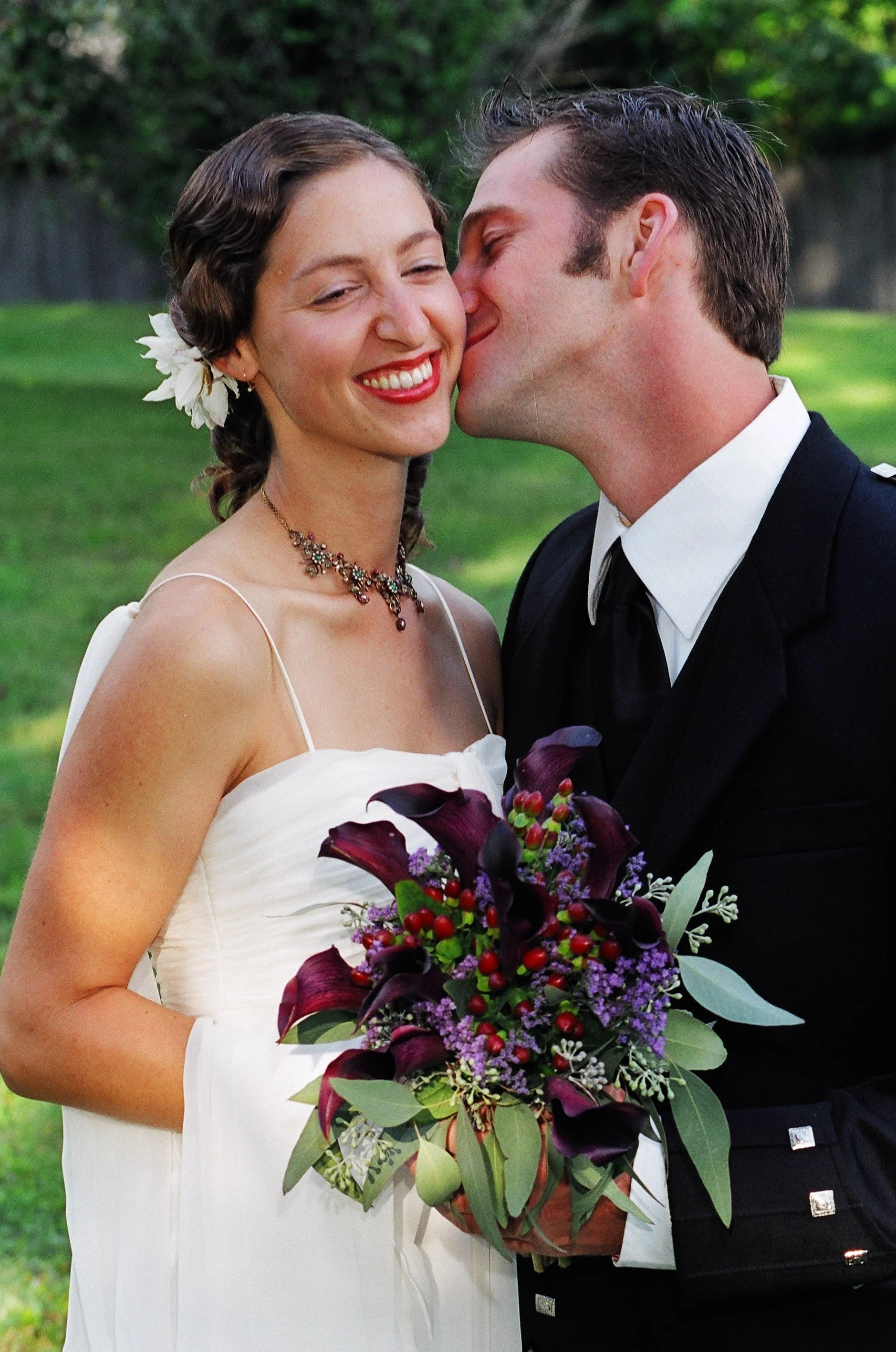 A bride and groom share a kiss outdoors, with the bride holding a floral bouquet, and both are smiling against a background of greenery.