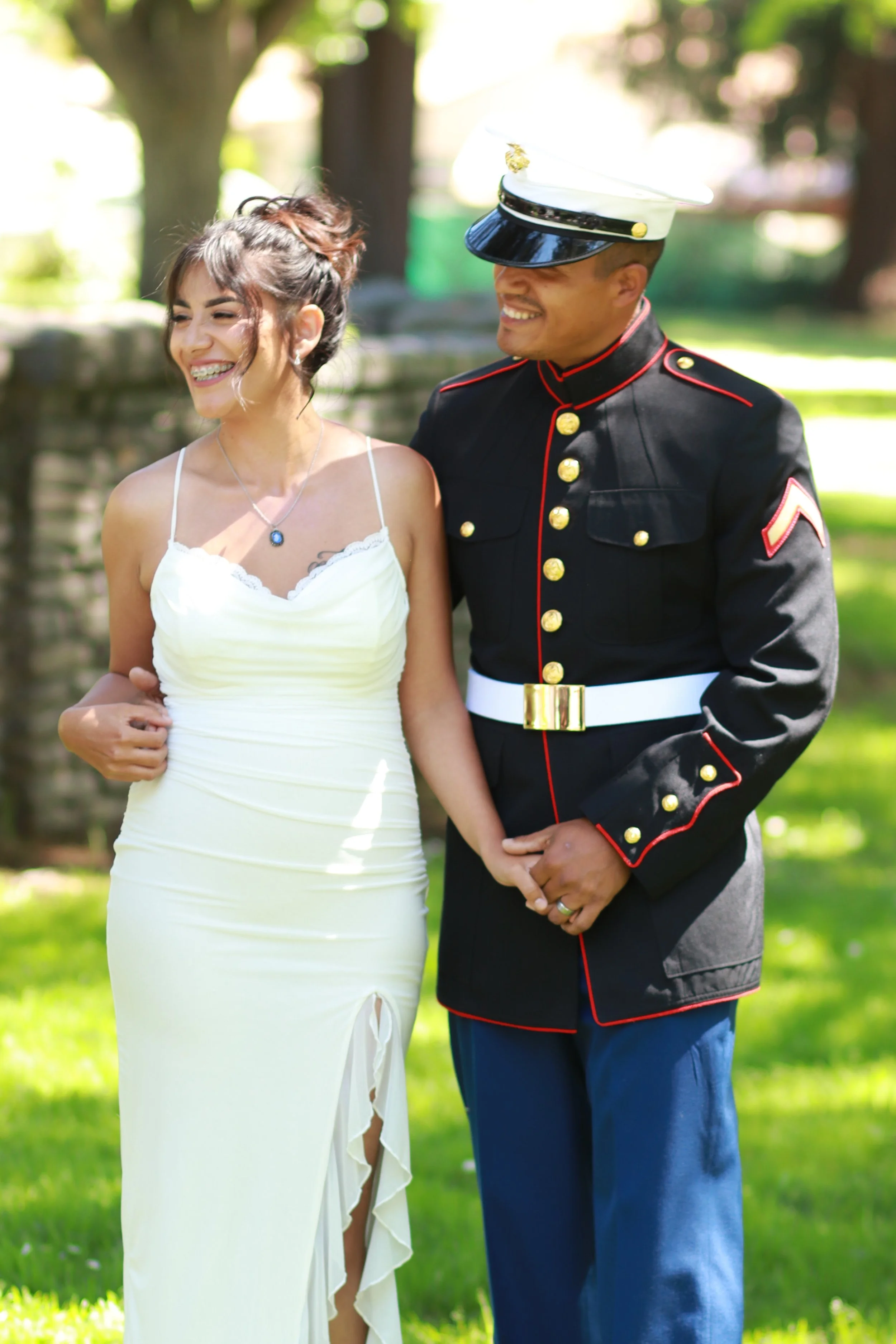A couple dressed in wedding attire, standing outdoors with trees and greenery in the background. The woman is wearing a white dress with a slit and a necklace, smiling with braces. The man is wearing a vintage military uniform with a white hat, gold 