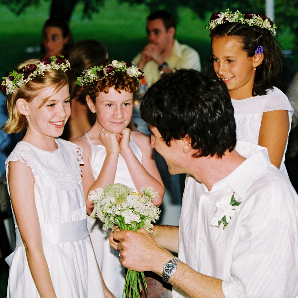 A man proposing marriage to a young girl holding a bouquet of flowers, with two other smiling girls watching. All are dressed in white and wearing flower crowns, outdoors at a celebration.