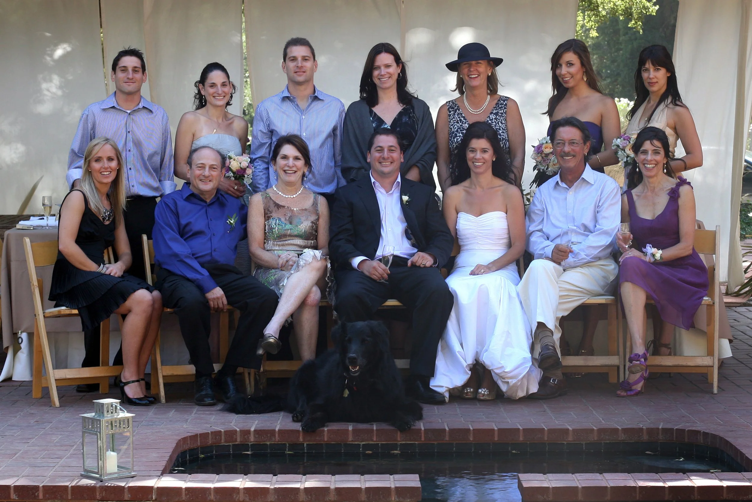 Group of people celebrating at a wedding, sitting and standing outdoors, with some holding bouquets and glasses, and a black dog sitting in front.