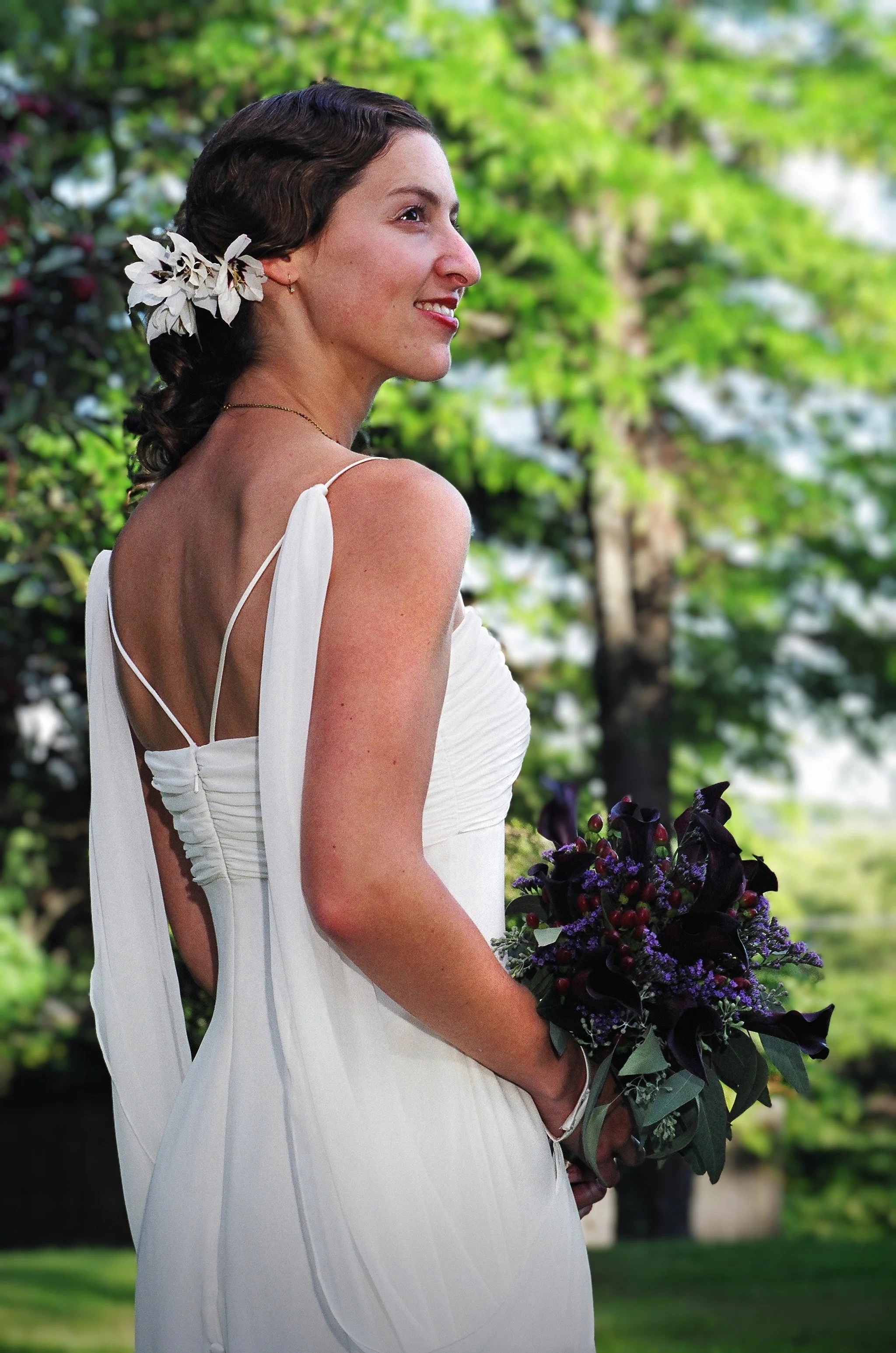A woman in a white dress with floral accents in her hair, holding a bouquet with dark purple and red flowers, outdoor with lush green trees in the background.