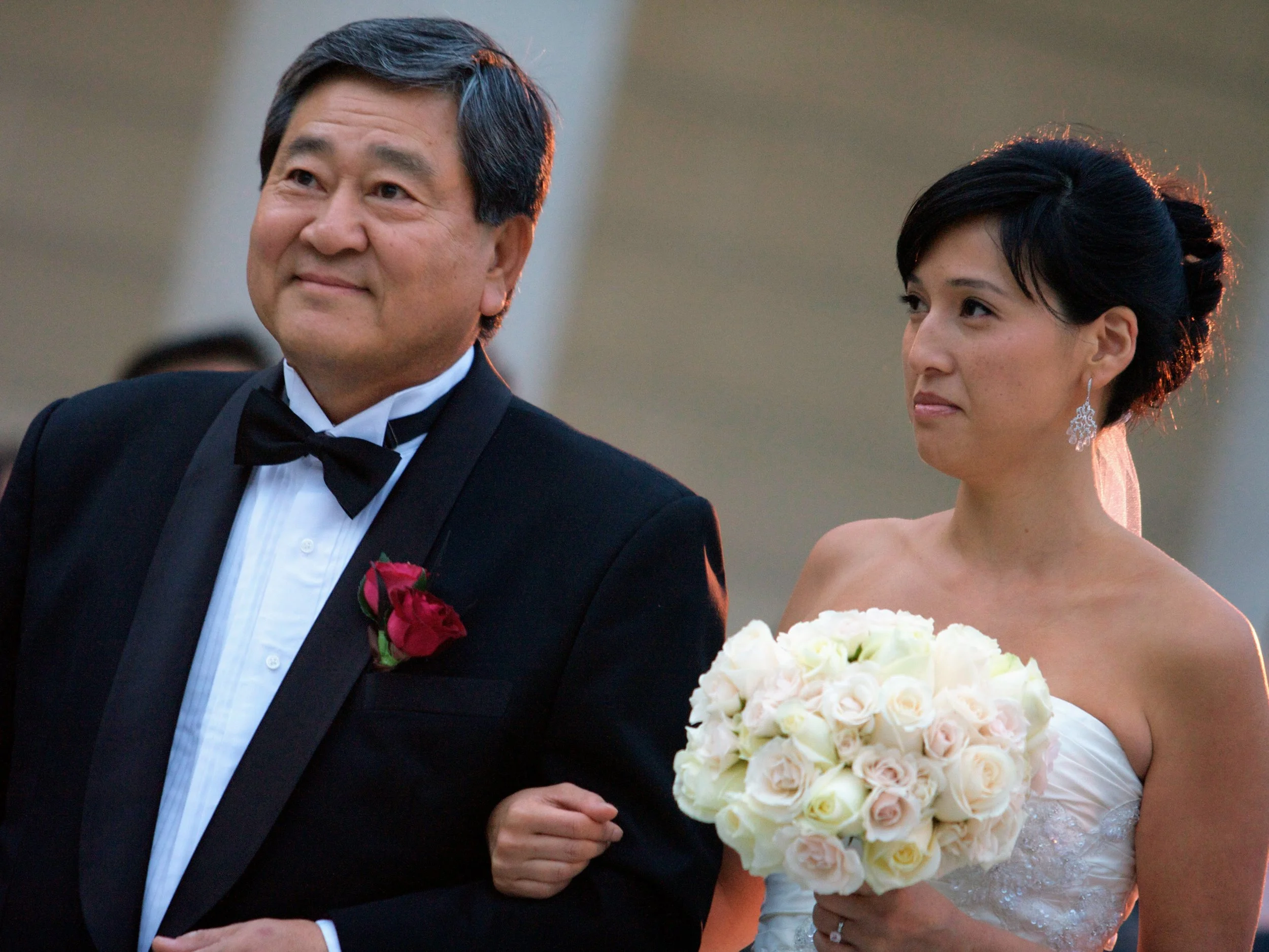 A man in a tuxedo and a woman in a wedding dress standing together, with the woman holding a bouquet of white and pink roses, at sunset outdoor wedding.