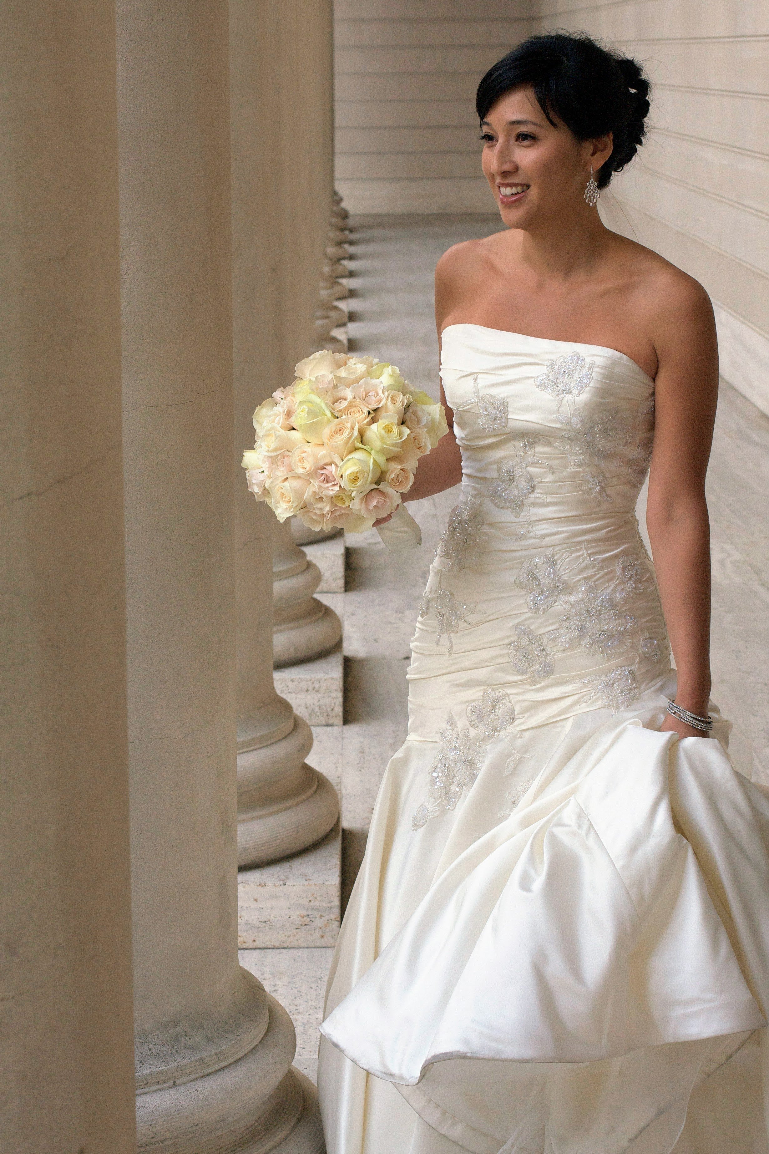 A woman in a white wedding dress holding a bouquet of cream and pink roses, smiling, standing in a corridor with stone columns.