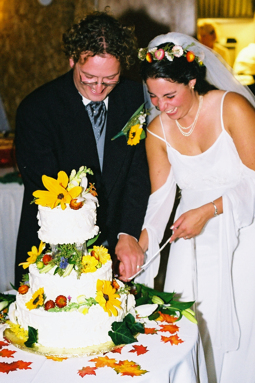 A bride and groom are cutting a three-tier wedding cake decorated with yellow and orange flowers and greenery at their wedding celebration.