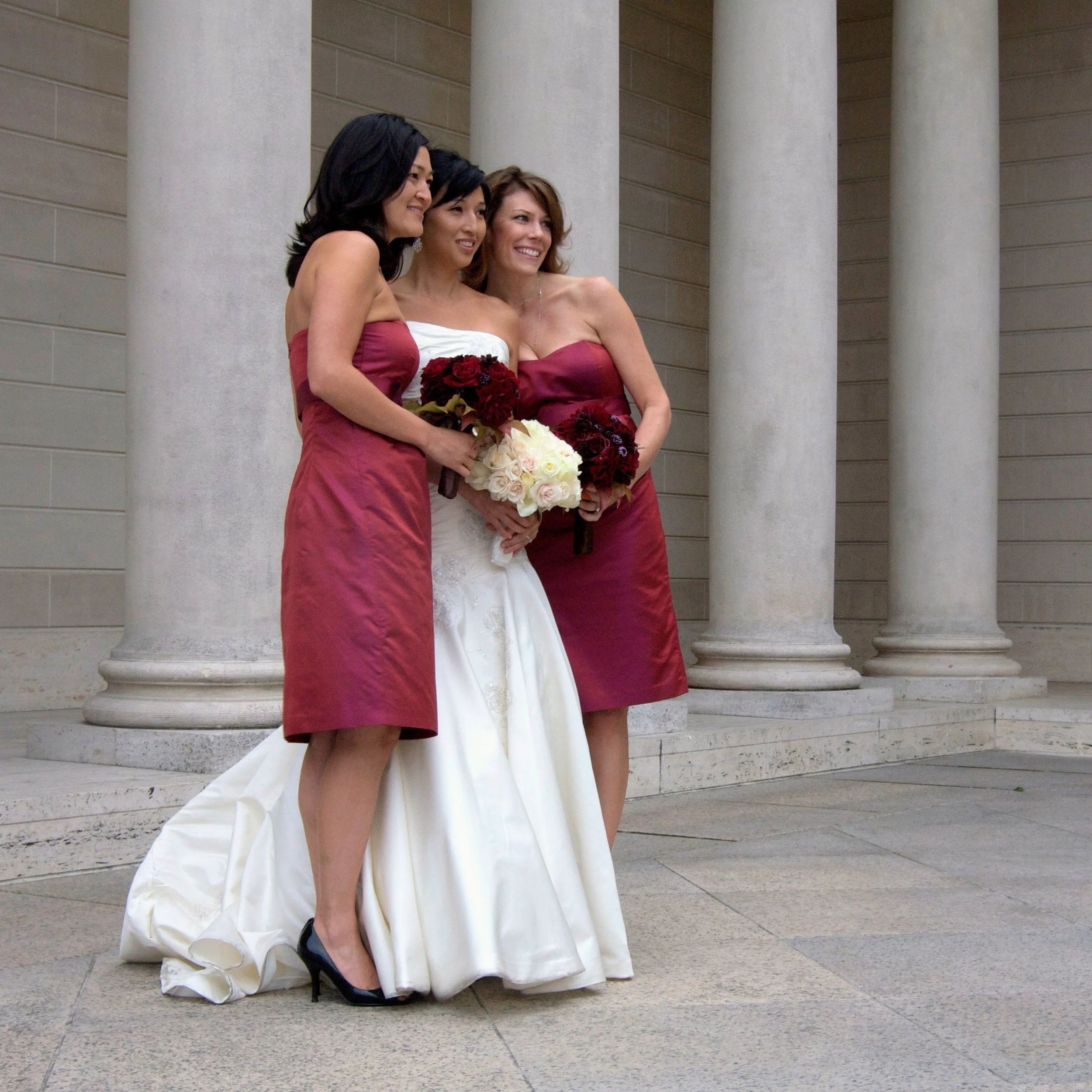 Three women in bridesmaid dresses standing together outside near stone columns, smiling and holding bouquets of red and white flowers.